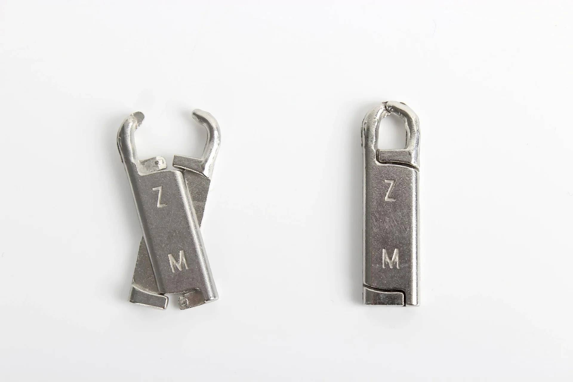 Zwei kleine Metallreißverschluss-Zuglasche oder Schiebeteile auf weißem Hintergrund, eines aufgebrochen und eines intakt, jeweils mit "Z"" und ""M"" gekennzeichnet.