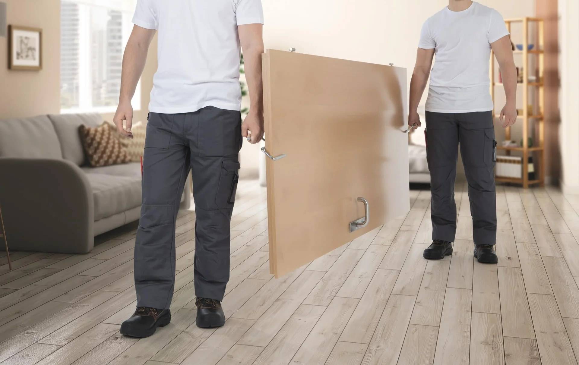 Zwei Männer in weißen Hemden und Arbeitshosen tragen ein großes Sperrholzbrett durch ein Wohnzimmer, was auf eine Hausrenovierung oder einen Umzug hindeutet.
