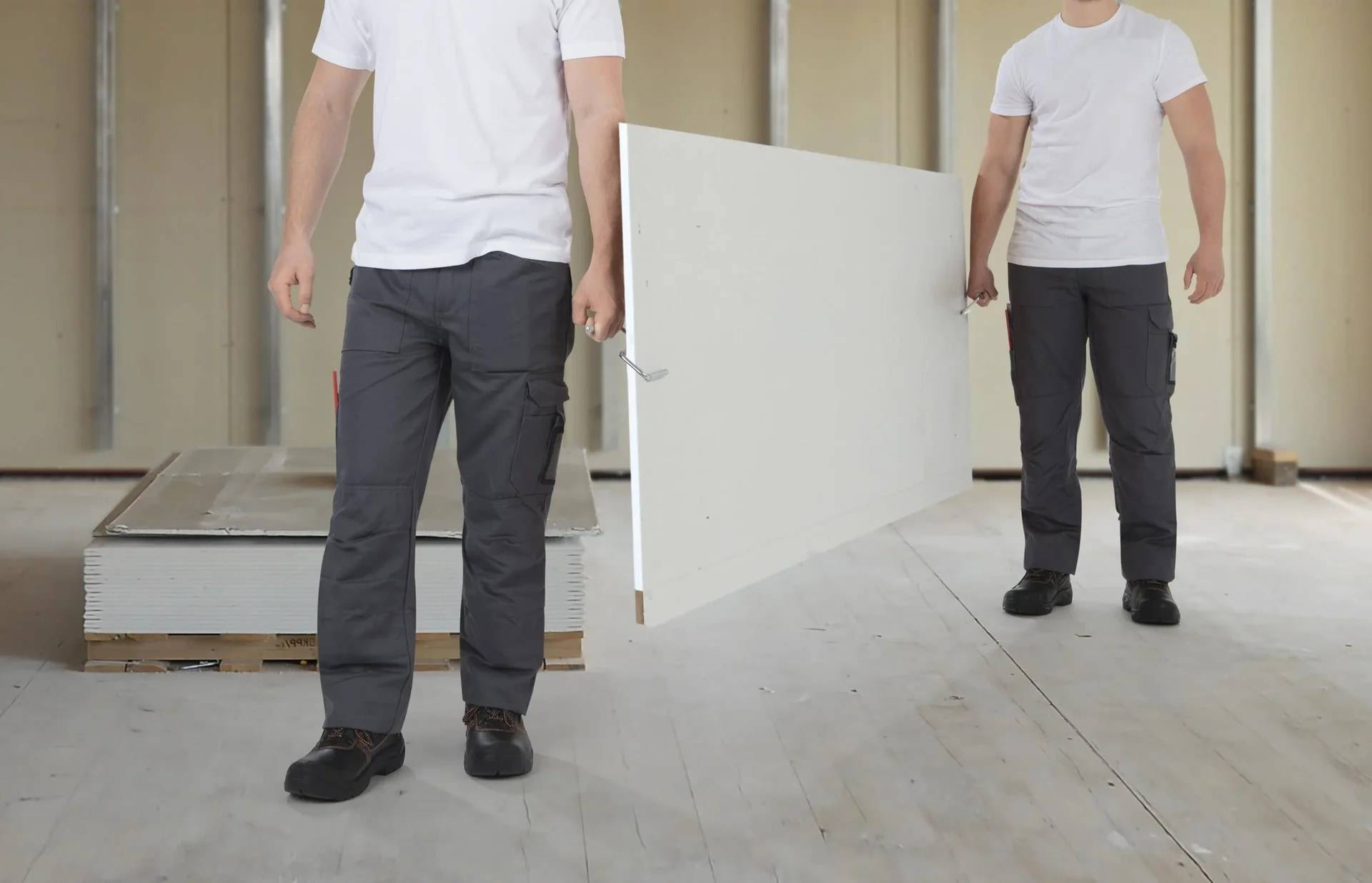 Zwei Bauarbeiter in weißen T-Shirts tragen ein großes weißes Panel durch einen unfertigen Innenraum.