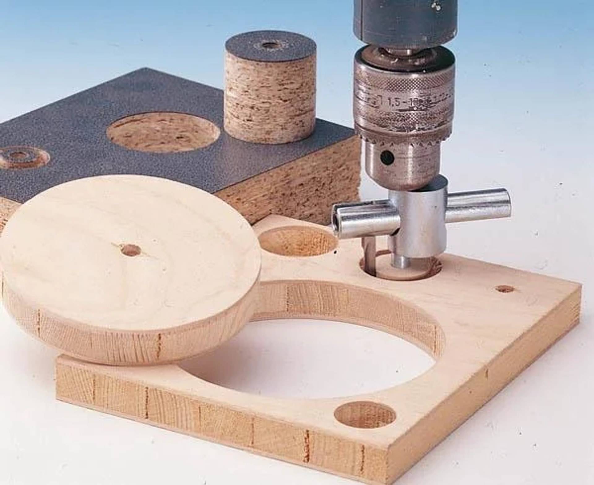 Eine Bohrmaschine schneidet kreisförmige Löcher in ein Holzbrett mit einer Lochsäge, wobei Verschnitte und eine große runde Holzplatte in der Nähe liegen.