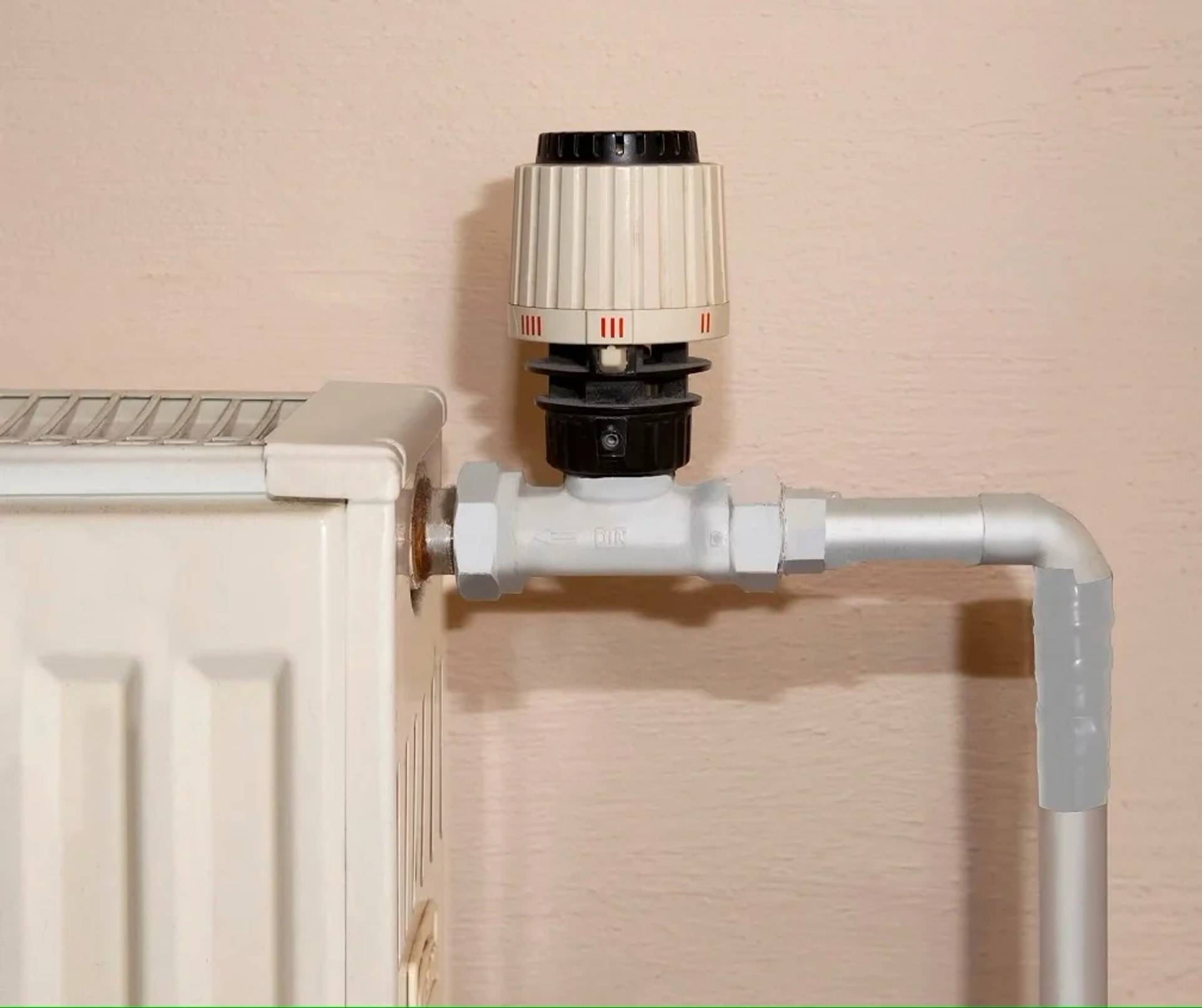 Ein Heizkörper mit thermostatischem Ventil und angeschlossenem weißem Heizungsrohr vor einer beigen Wand.