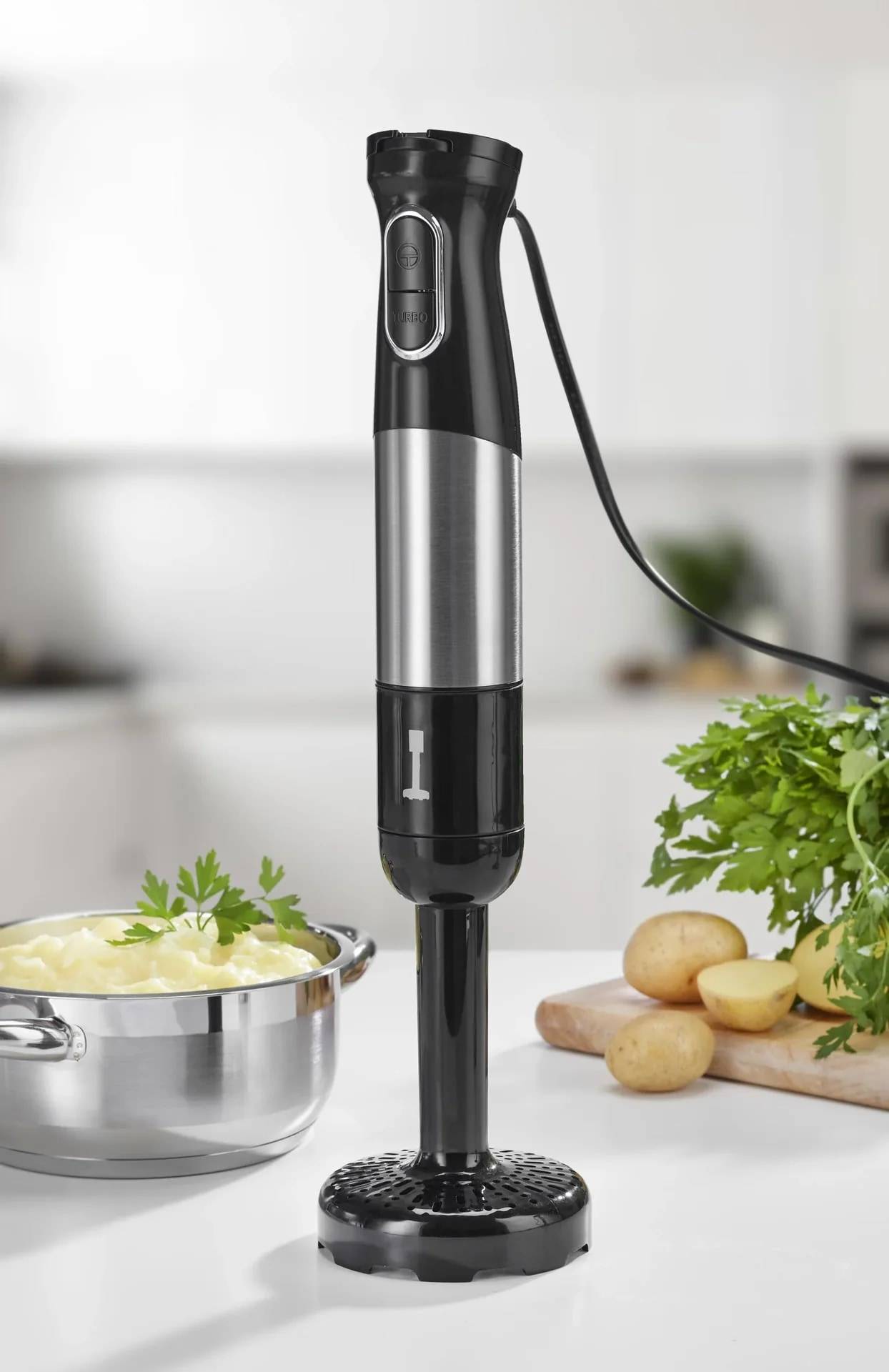 Schwarz-silberner Stabmixer, der in einer Küche neben einem Topf Suppe, Kartoffeln und Petersilie auf einer Arbeitsplatte steht.