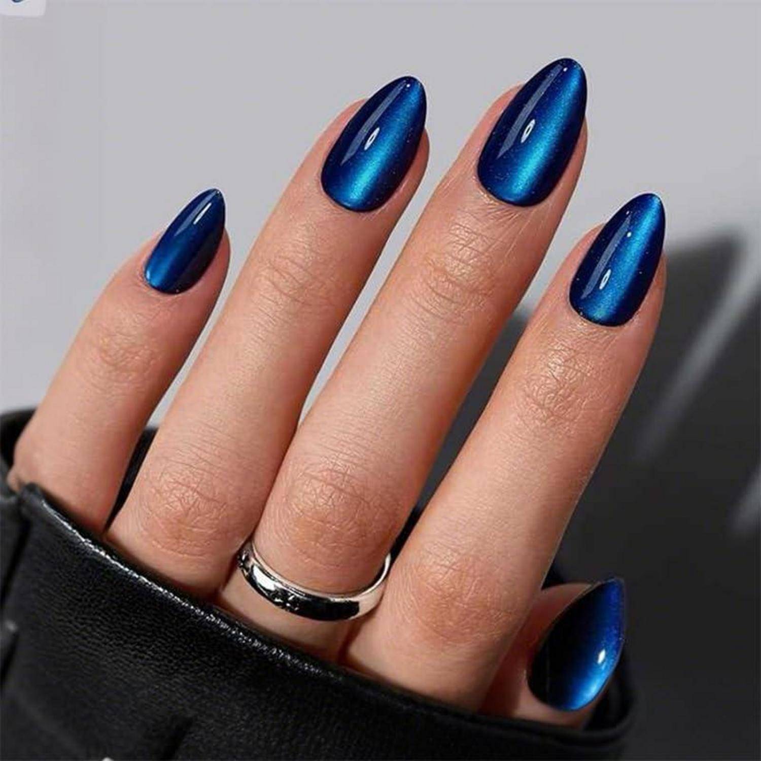 Nahaufnahme einer Hand mit langen metallisch blauen mandelförmigen Nägeln und einem silbernen Ring, der aus einem schwarzen Handschuh vor grauem Hintergrund hervorragt.