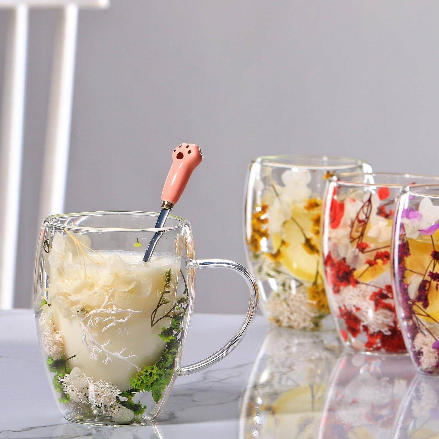 Glastasse mit Blütentee und einem Löffel mit rosa Griff, gezeigt zusammen mit drei weiteren durchsichtigen Tassen mit farbenfrohen blütengetränkten Getränken auf einer reflektierenden Oberfläche.
