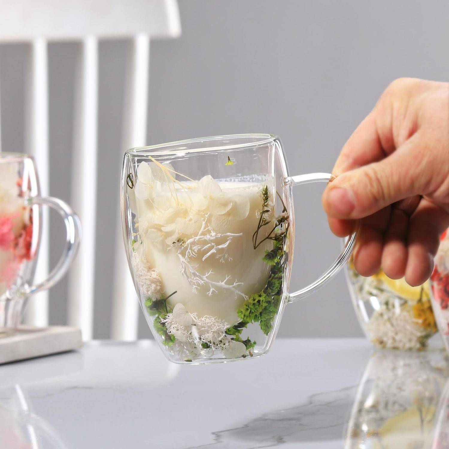 Hand, die eine klare Glastasse mit einer weißen Blumenarrangement und Grünzeug innen hält, gezeigt auf einer Tischplatte mit anderen unscharfen Blumenbechern im Hintergrund.