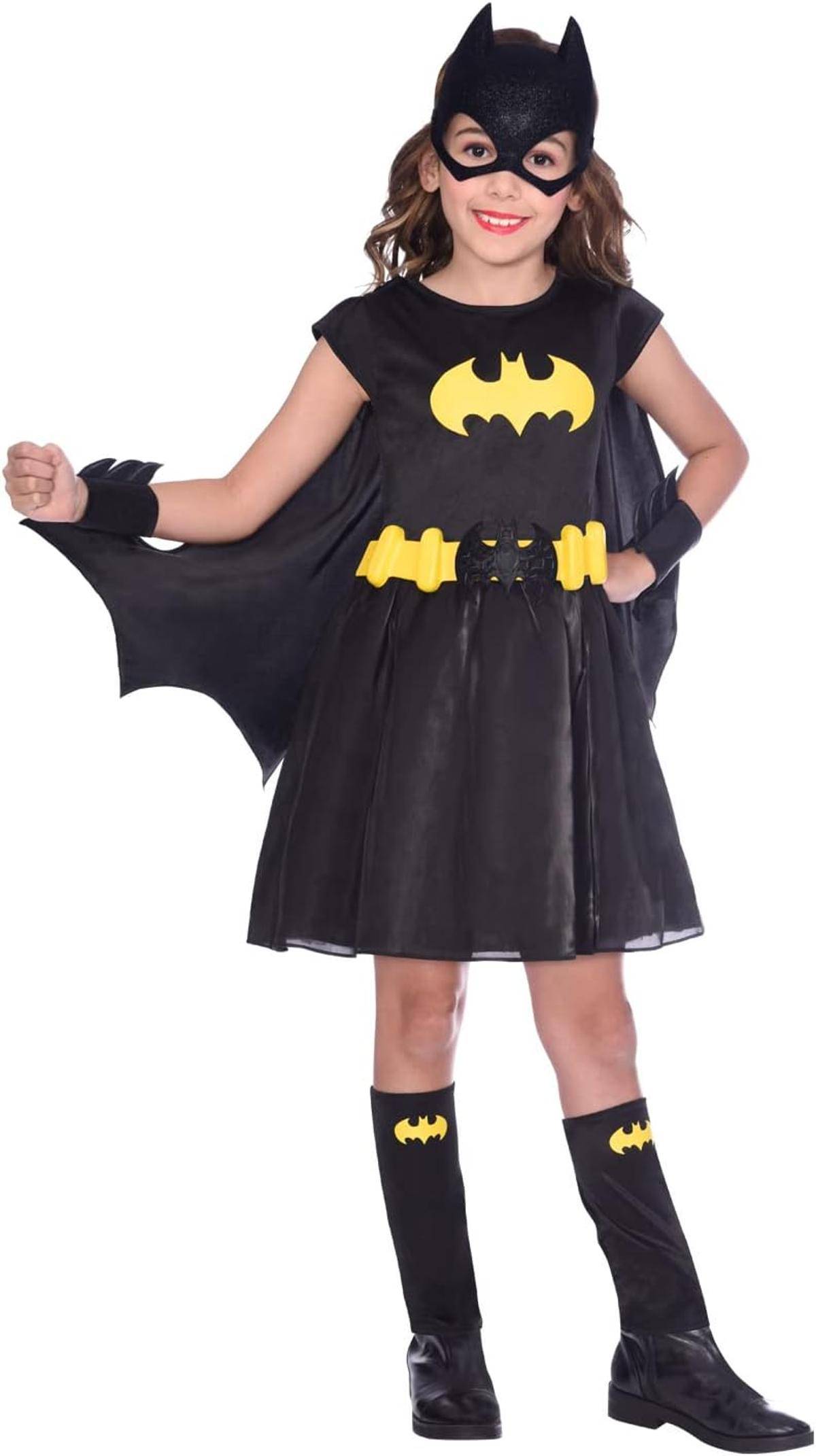 Junges Mädchen in einem Batgirl-Kostüm, das eine schwarze Maske, einen Umhang, ein Kleid, einen Gürtel und Stiefel trägt, posierend vor einem einfachen weißen Hintergrund.