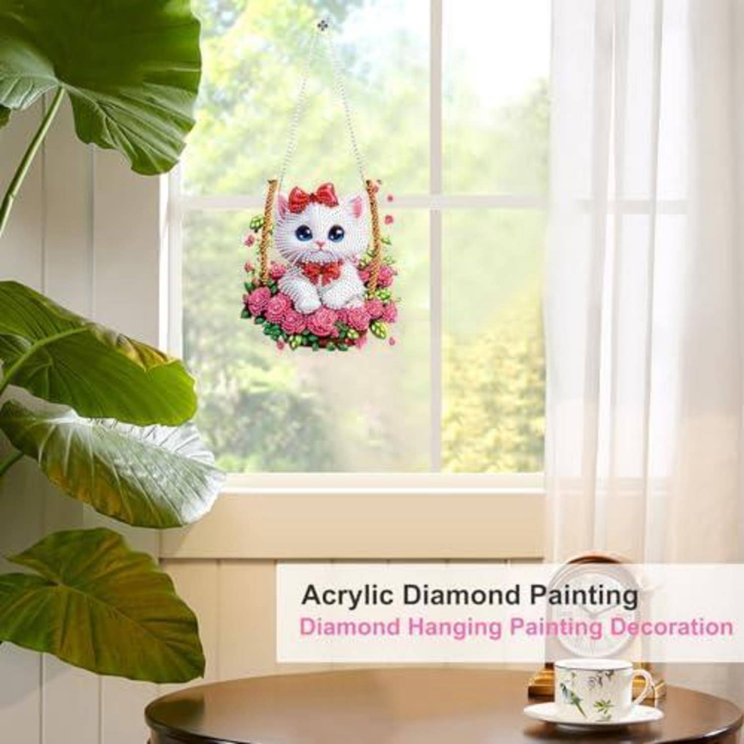Eine süße, katzenförmige Diamantmalerei-Hängdekoration mit rosa Blumen, die von einem sonnigen Fenster über einem Tisch und einer Teetasse angezeigt wird.