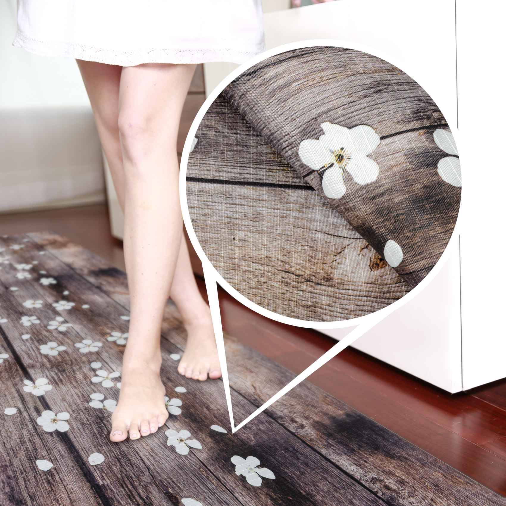 Nackte Füße neben einer Fußmatte mit Blumenmuster auf einem Boden, mit vergrößerter Detailansicht, die die Holzmaserung der Matte und das weiße Blumendesign zeigt.