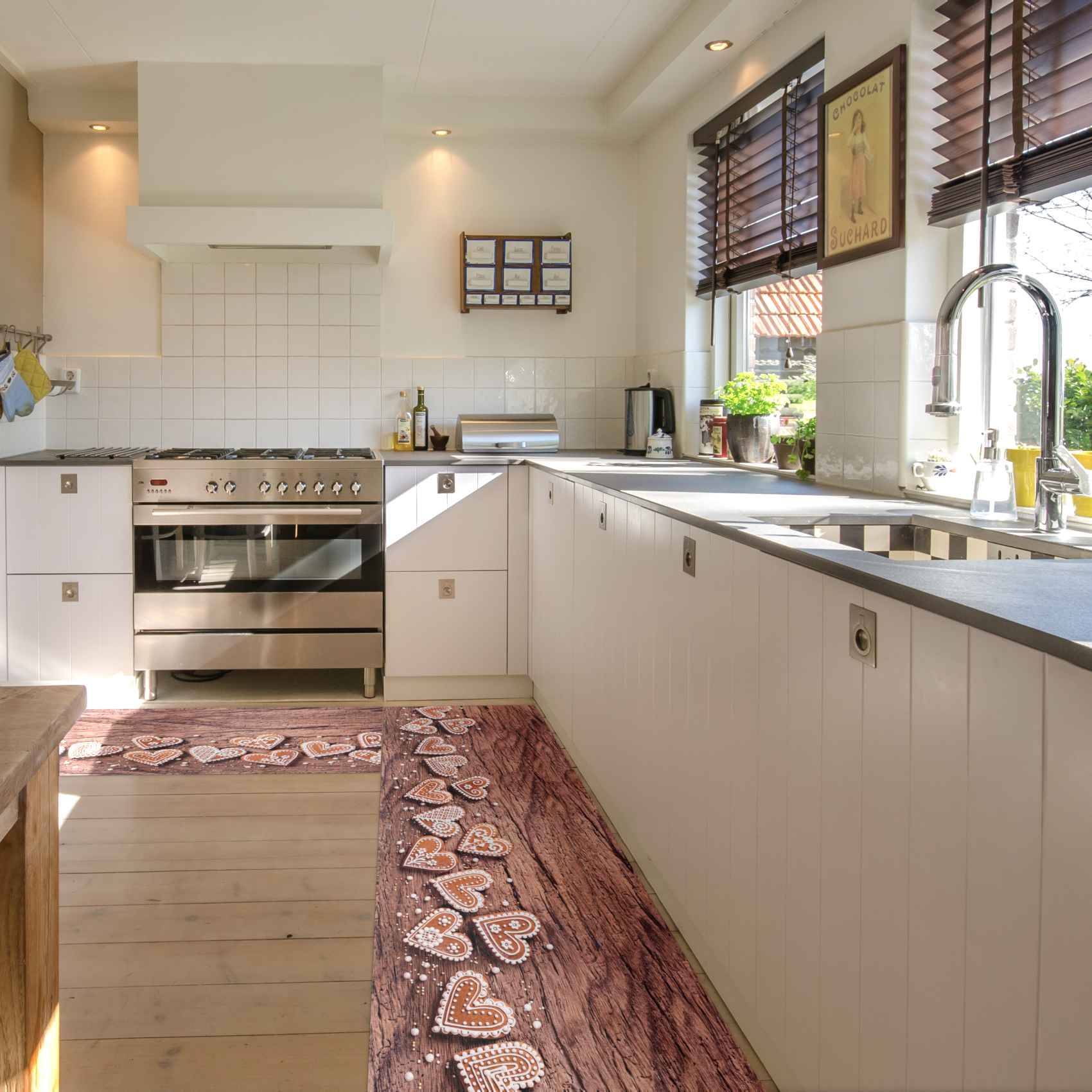 Helle, moderne Küche mit weißen Schränken, Edelstahl-Backofen, langer Arbeitsplatte, Spüle am Fenster und gemusterten Teppichen auf Holzboden.