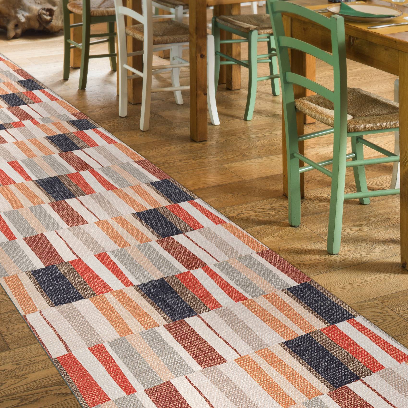 Farbenfroher geometrischer Läufer mit roten, dunkelblauen, beigen und orangefarbenen Blöcken auf einem Holzboden neben nicht zusammenpassenden Esszimmerstühlen und einem Tisch.