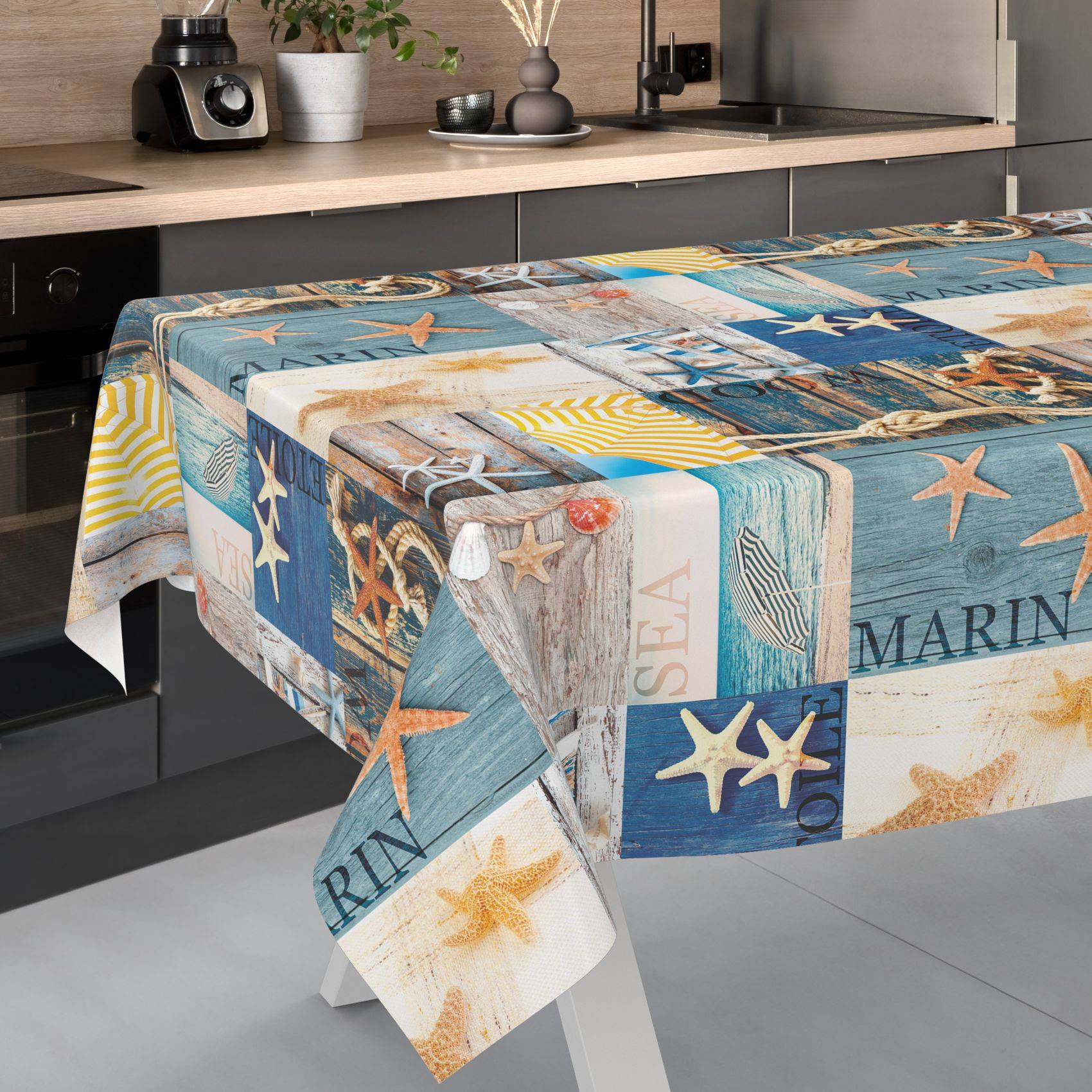 Nahaufnahme eines Tisches, der mit einer maritim gestalteten Tischdecke bedeckt ist, die Muscheln, Seesterne, Anker und Meerestext in Blau, Beige und Weiß zeigt.