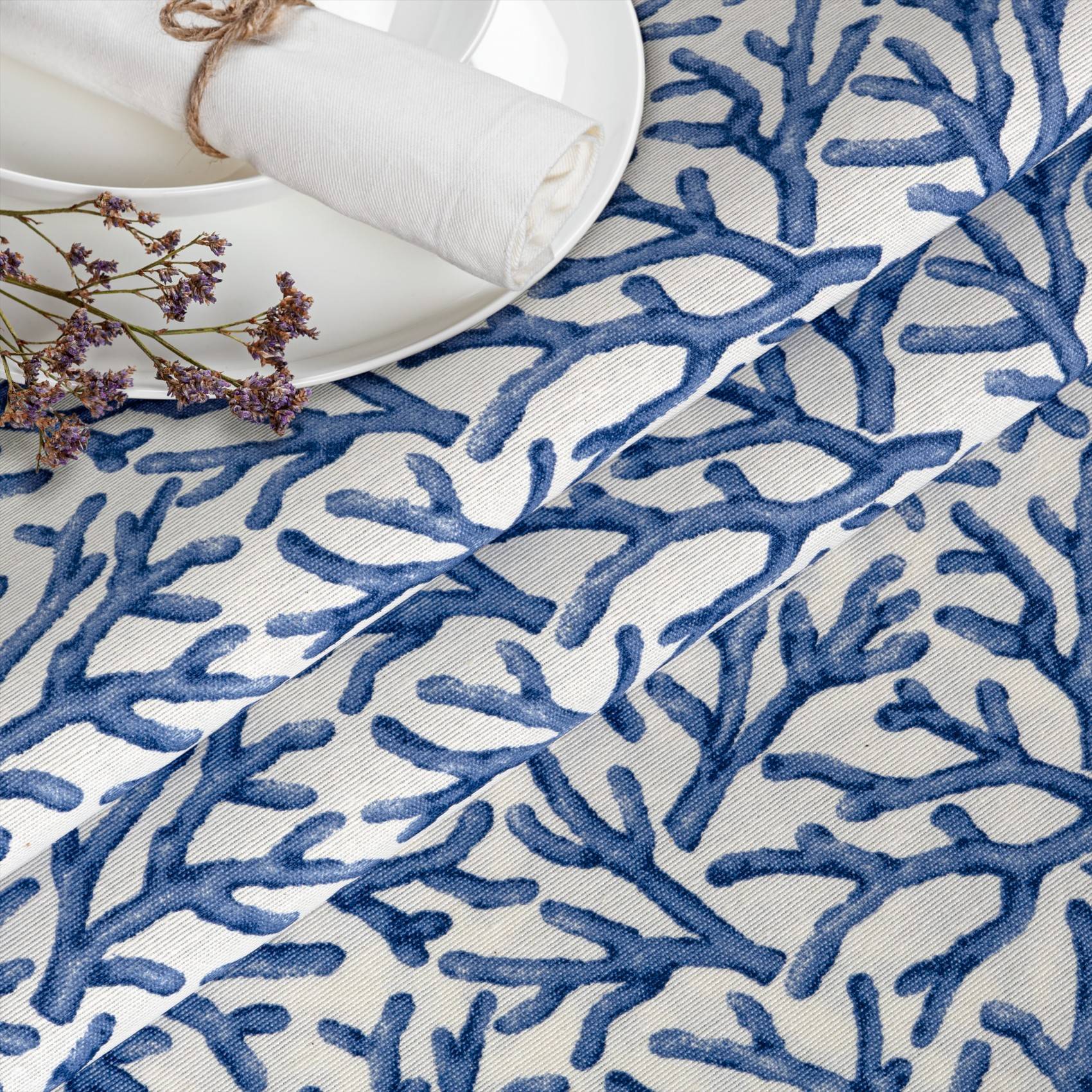 Blaue Tischdecke mit Korallenmuster mit weißem Teller, zusammengerollter Serviette und Trockenblumen in der Ecke