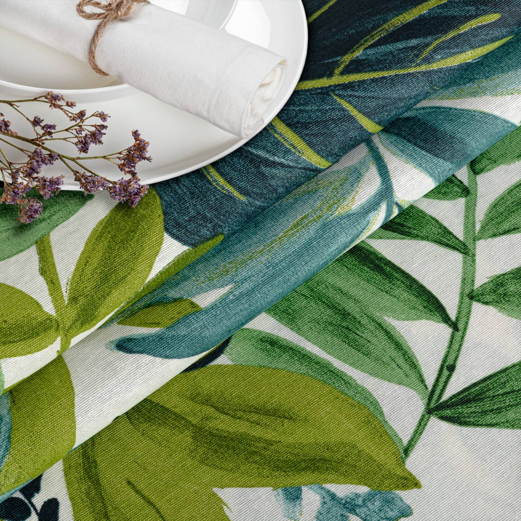 Gemusterter grün und blau bedruckter Stoff mit Blattmotiv, eine gefaltete Tischdeckung und kleine violette Blüten in der Ecke.