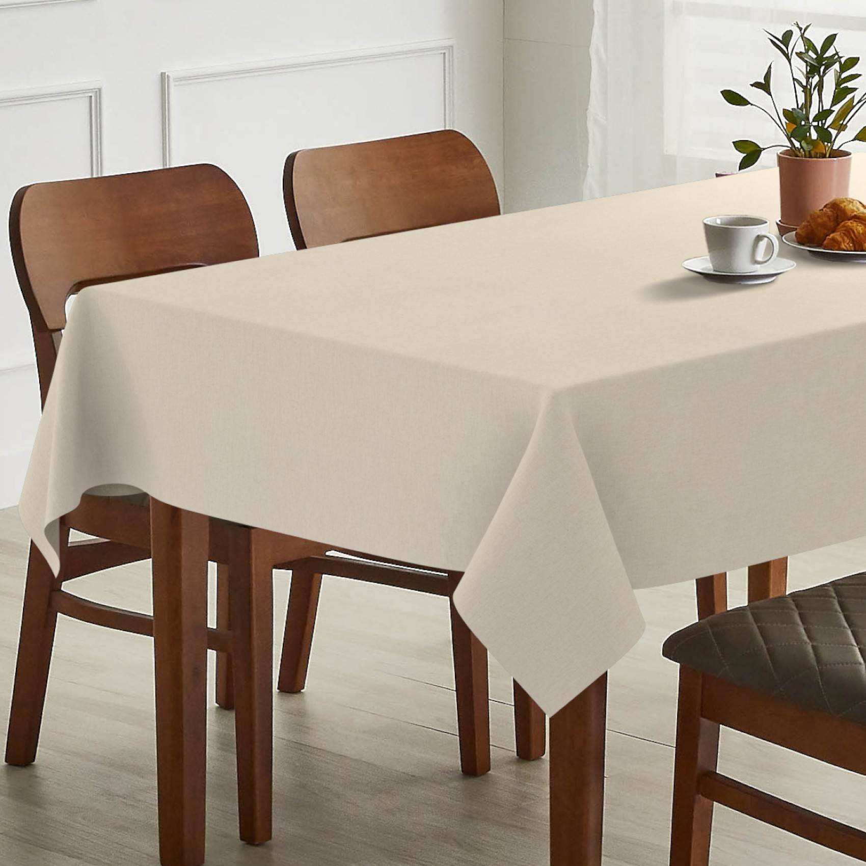 Minimalistisches Esstisch-Setting mit beiger Tischdecke, hölzernen Stühlen, einer Kaffeetasse, einem Croissant und einer Topfpflanze in einem hellen Raum.