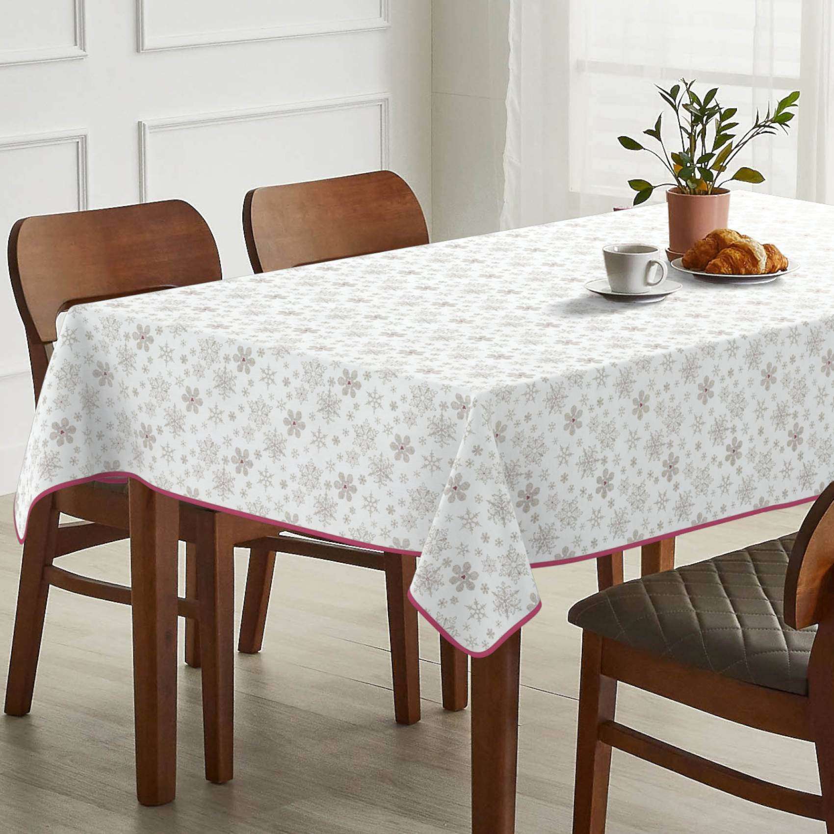 Esstisch mit einer weißen Tischdecke mit Blumenmuster und rosa Borte, eingedeckt mit Kaffee, Croissants und einer Topfpflanze in einem hellen Raum mit Holzstühlen.