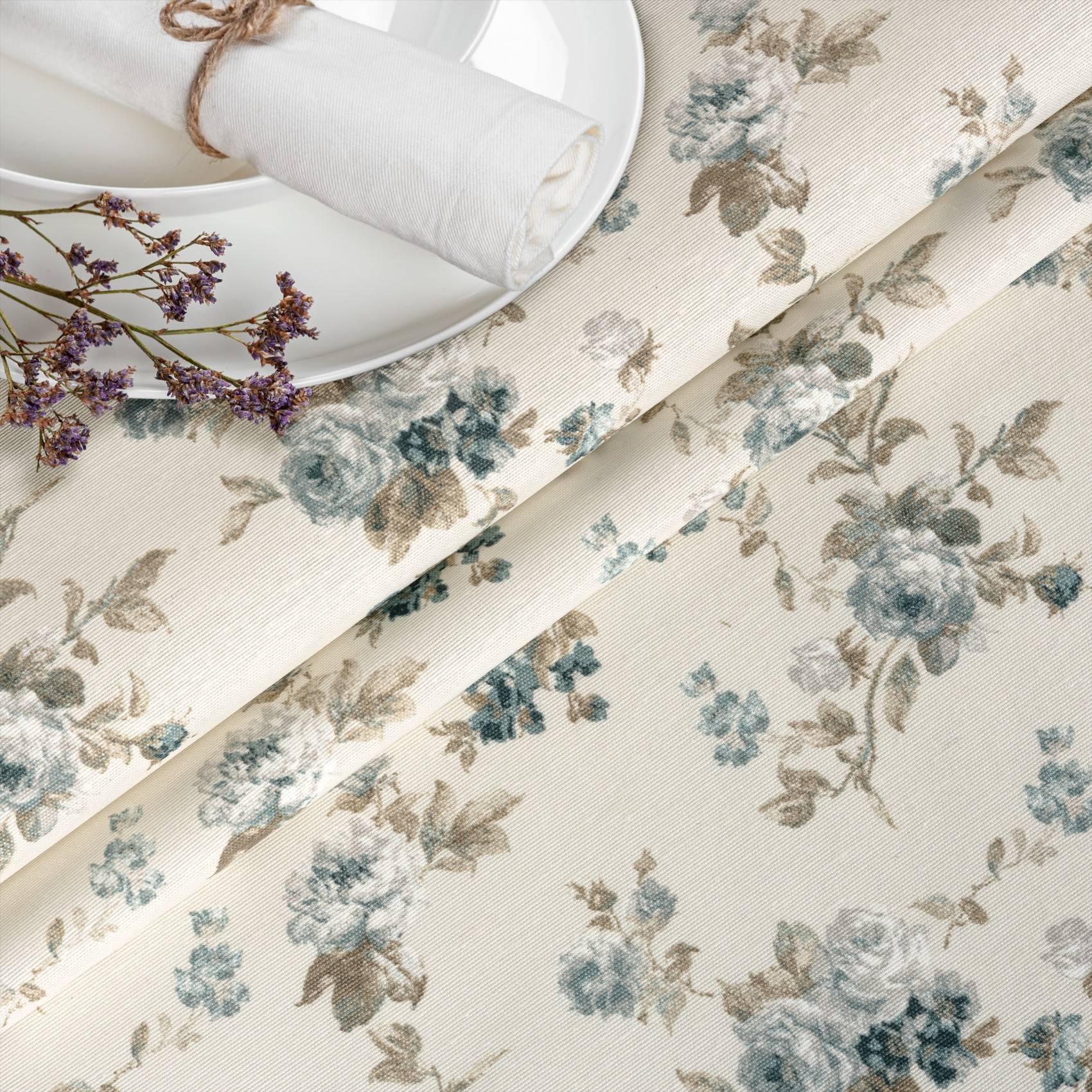 Blumenmusterter Stoff in zartem Blau und Beige mit einer gefalteten weißen Serviette und einem Teller, was eine Tischdecke oder Heimtexstil andeutet.
