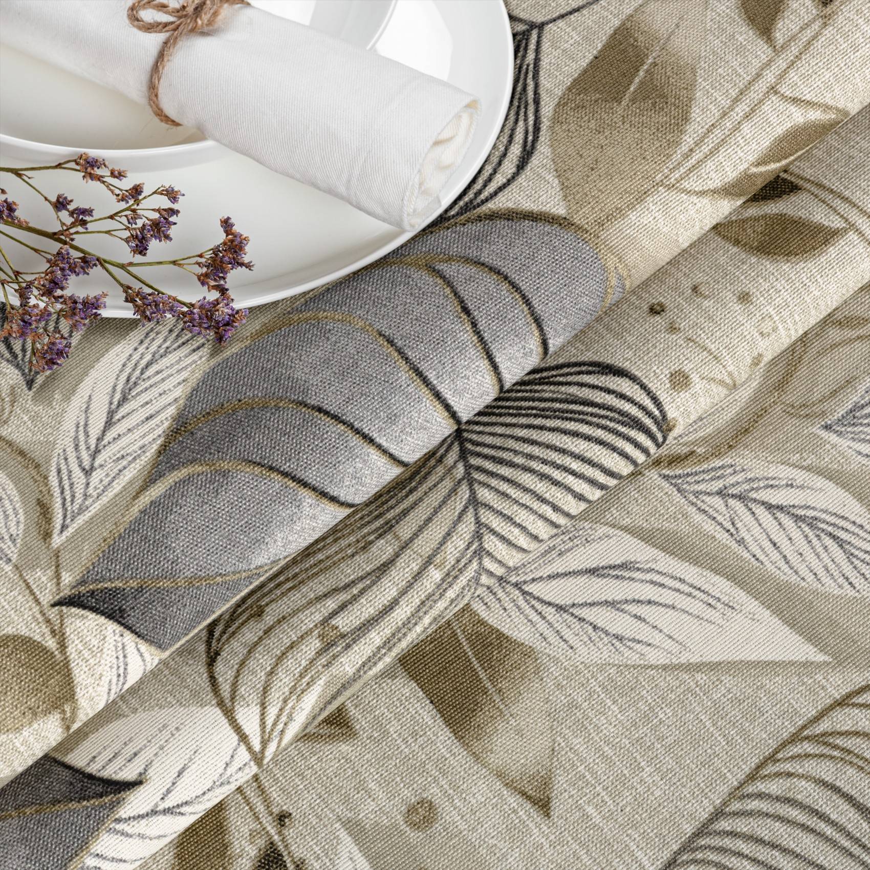 Nahaufnahme von gerolltem gemustertem Stoff mit Blatt- und Astdrucken in Beige, Grau und Schwarz, dargestellt neben einem weißen Teller und getrockneten Blumen.