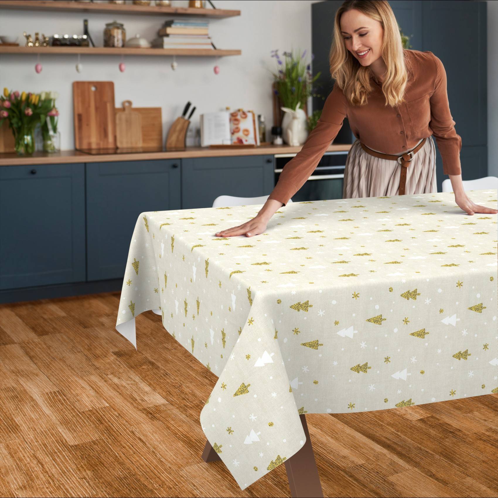 Eine Frau glättet eine festliche weiße Tischdecke mit gold- und silbernen Weihnachtsbäumen und Sternenmustern über einen Esstisch in einer Küche.
