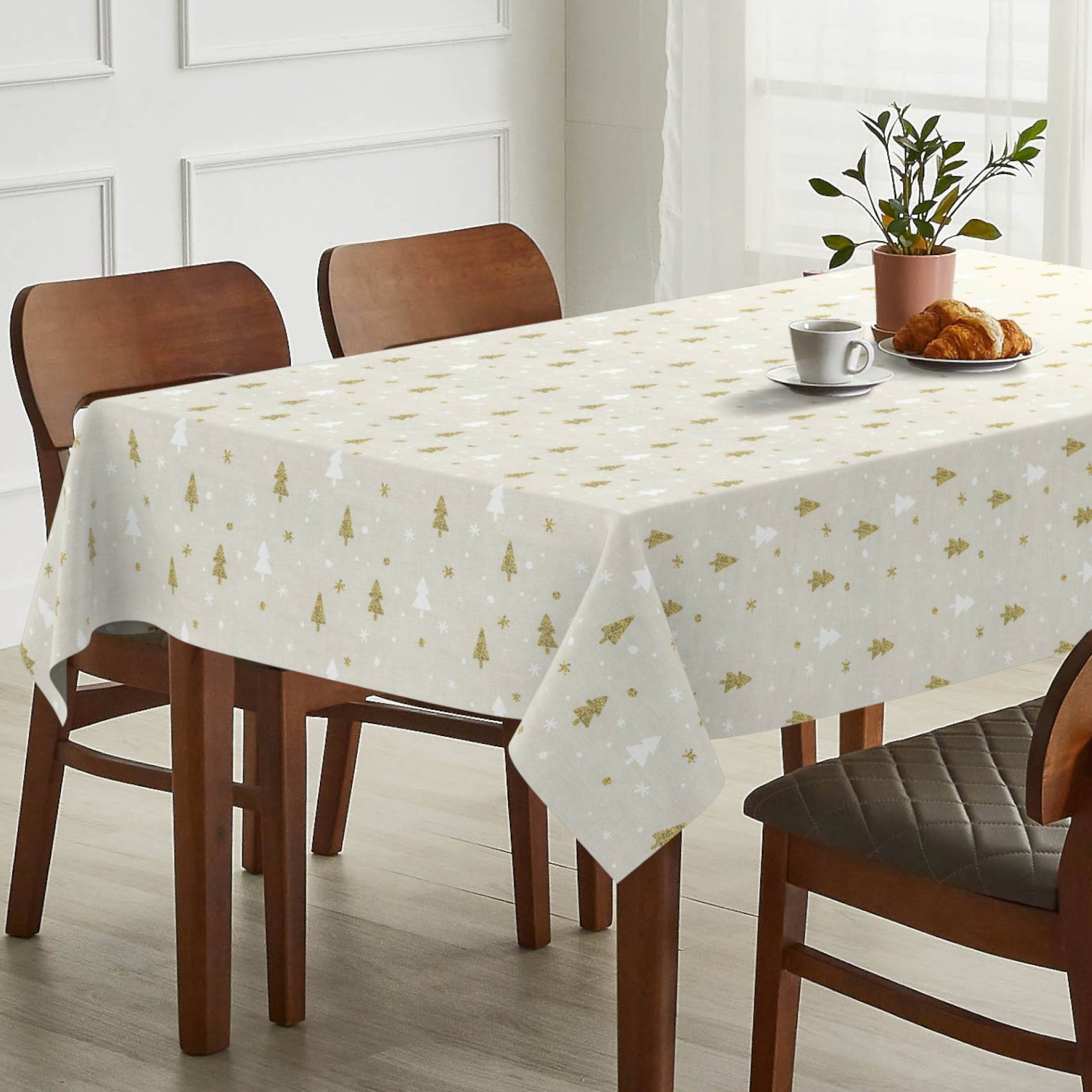 Esstisch mit einer weißen Weihnachtsmuster-Tischdecke, einer Kaffeetasse, einem Croissant-Teller und einer Topfpflanze in einem hellen Zimmer mit Holzstühlen.