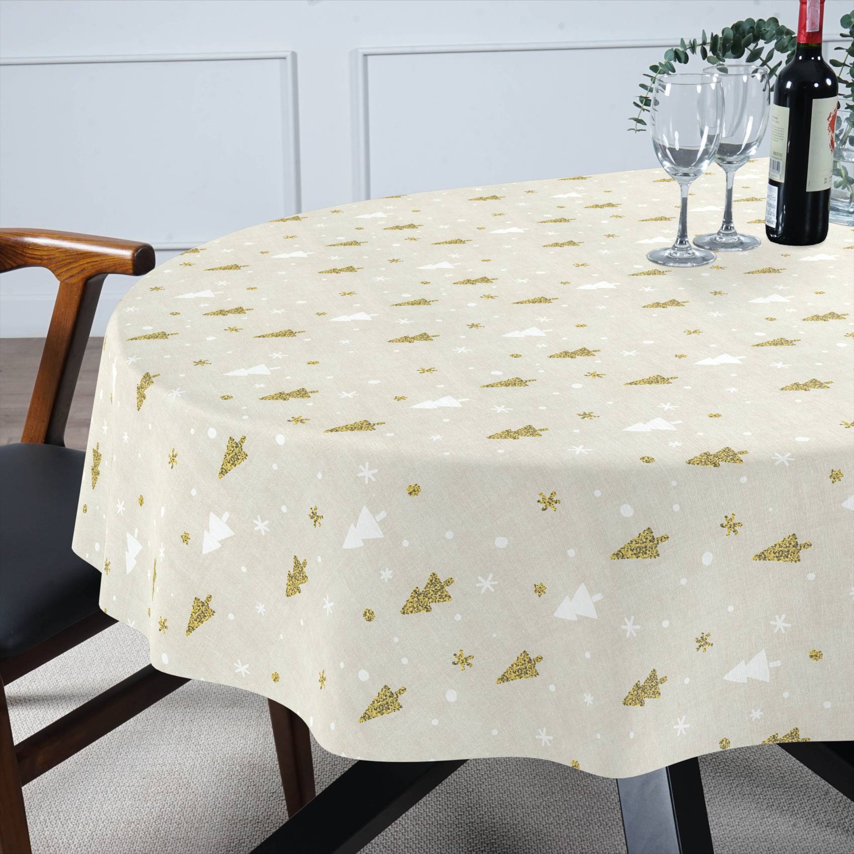 Rundes Esstisch bedeckt mit einer beige Tischdecke mit Gold- und Weihnachtsbaummustern in Gold und Weiß sowie Schneeflockenmustern, eingedeckt mit Weingläsern und einer Flasche.