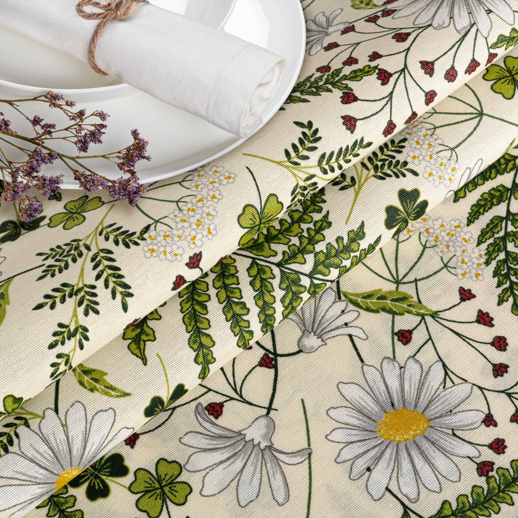 Blumige Tischdecke mit weißen Gänseblümchen, Klee und Grünwerk, gezeigt mit einem weißen Teller und einer aufgerollten Serviette auf einem Esstisch.