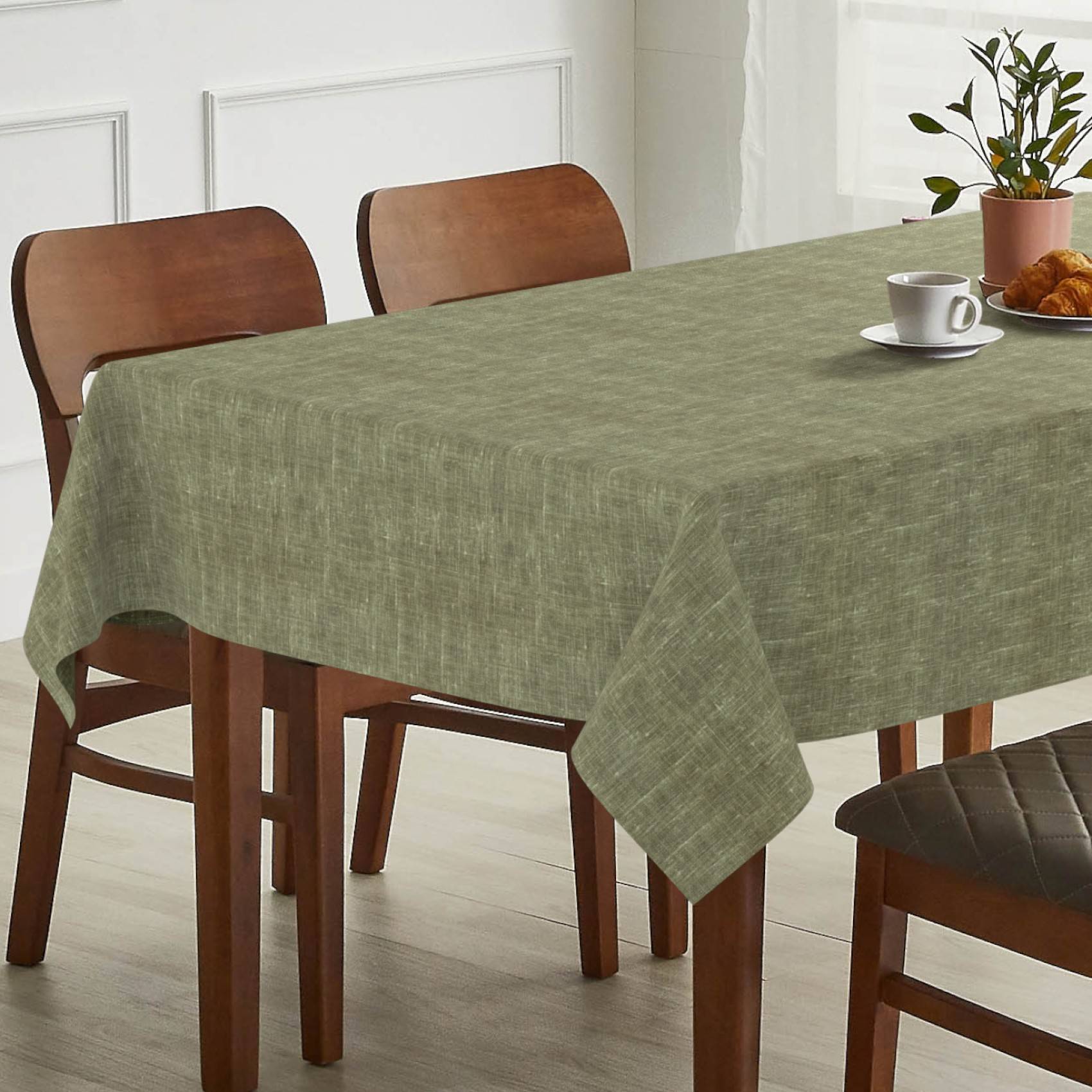 Esstisch mit grüner Tischdecke, zwei Holzstühlen und einer Tasse Kaffee mit Croissant neben einer Topfpflanze.