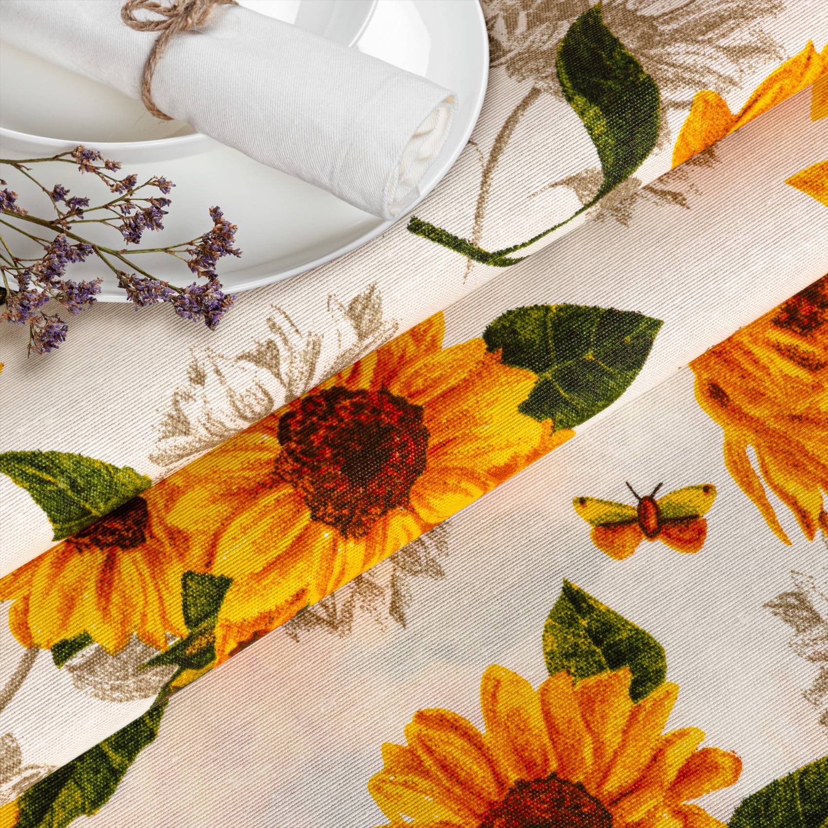 Tischdecke oder Stoff mit Sonnenblumenmuster oder Schmetterlingsmotiv, gezeigt neben einem weißen Teller, einer gefalteten Serviette und getrockneten Lavendelzweigen.