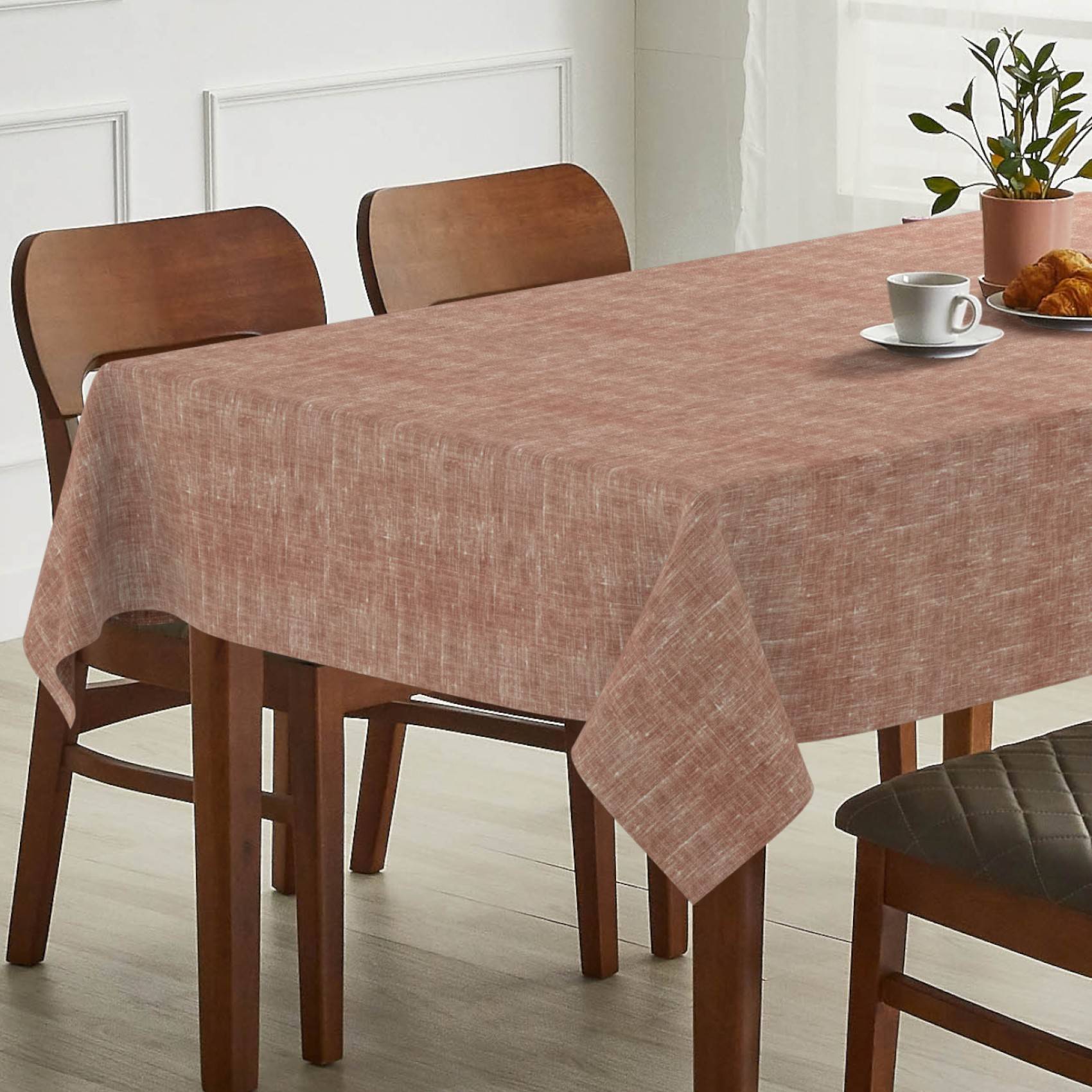 Esstisch mit einer staubig rosa Tischdecke, Holzstühlen und einer Tasse, Untertasse, einem Croissant und einer Topfpflanze auf einem hellen Boden.