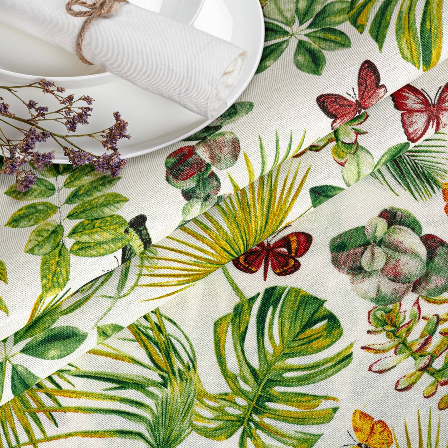 Bunter botanischer Tischdecke mit tropischen Blättern und Schmetterlingen, neben weißem Geschirr und einer aufgerollten Serviette auf einem gedeckten Tisch.