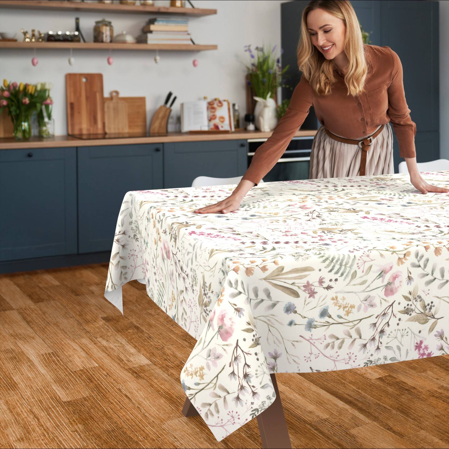 Eine Frau glättet eine gemusterte Tischdecke über einem Esstisch in einer Küche mit blauen Schränken und Holzboden.