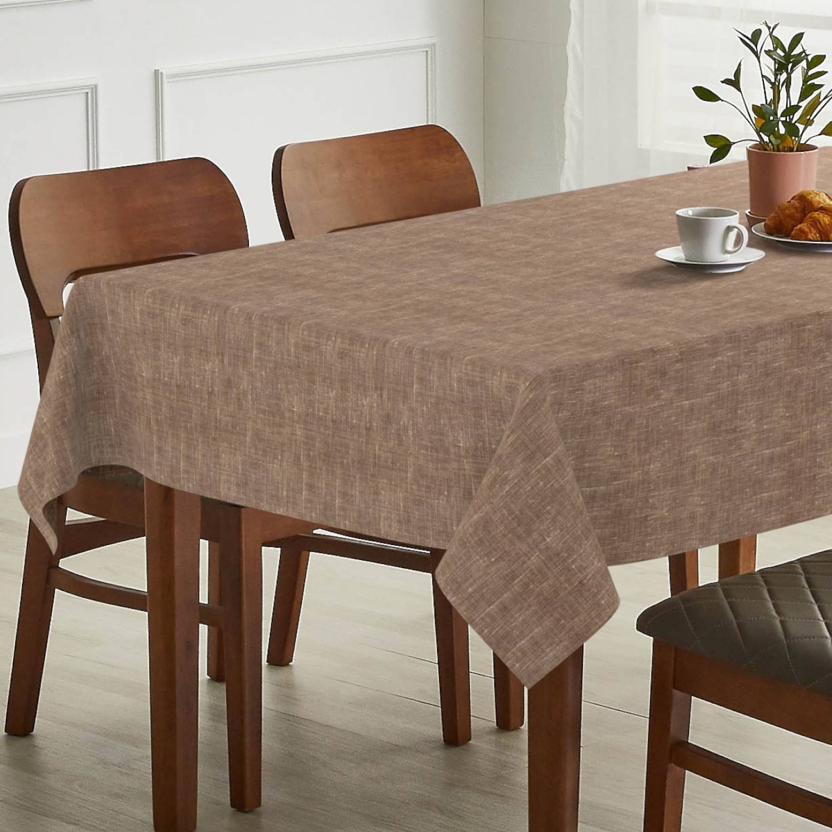 Esstisch mit brauner Tischdecke, Holzstühlen und einer Tasse mit Croissant sowie einer Topfpflanze auf der Tischplatte.