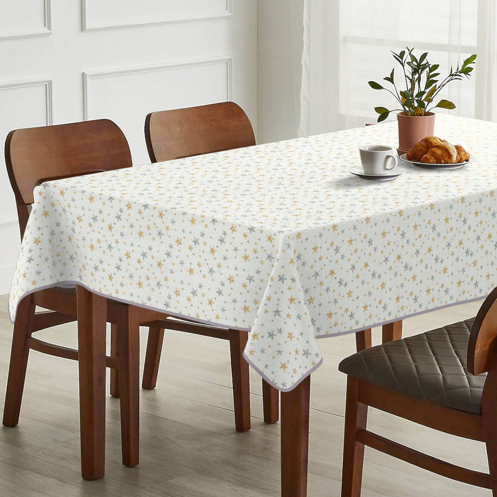 Esstisch mit einer sternengemusterten Tischdecke, gedeckt mit einer Tasse, einem Croissant-Teller und einer Topfpflanze in einem hellen Raum mit Holzstühlen.