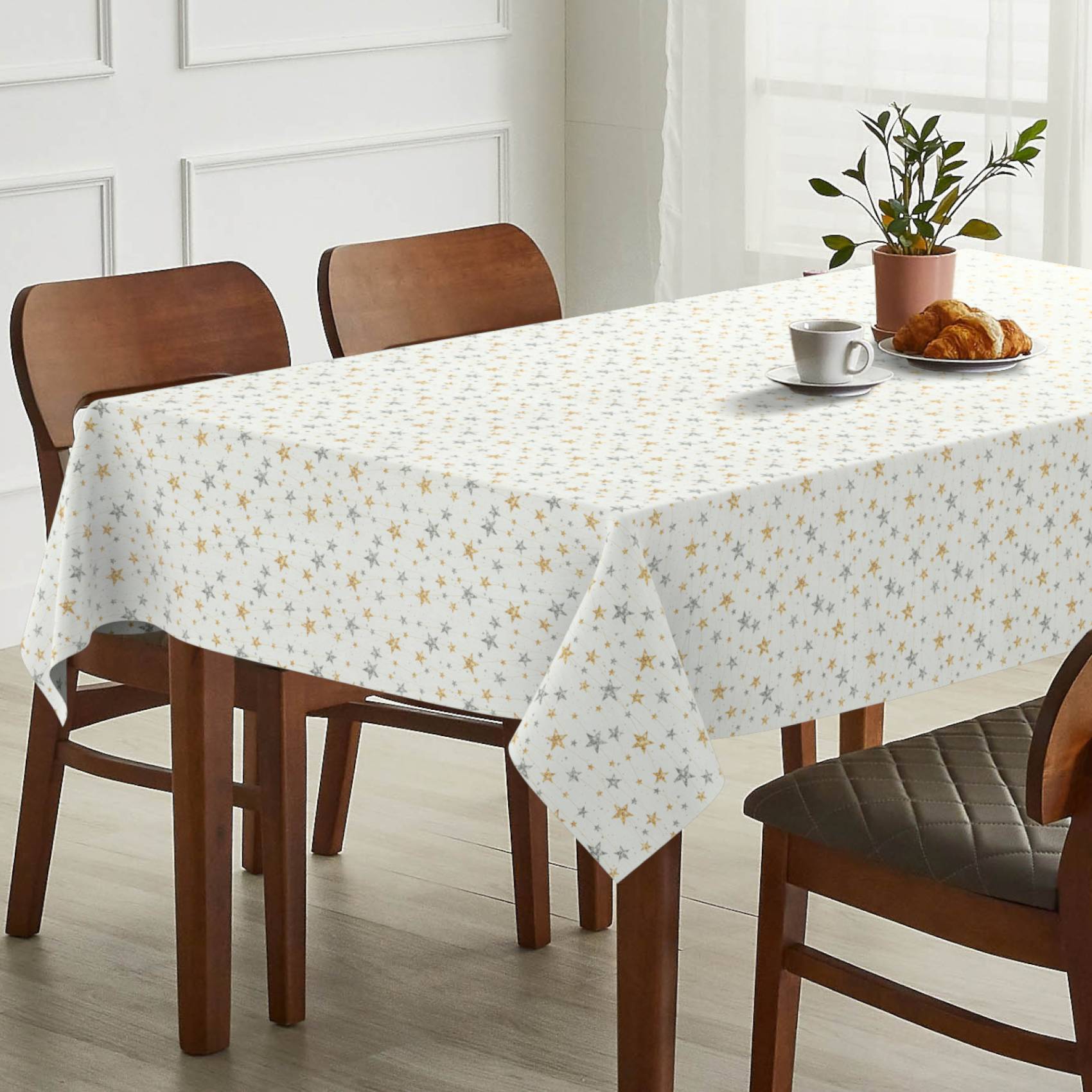 Esstisch mit weißer Tischdecke mit Sternenmustern, eingedeckt mit einer Tasse, Croissant-Teller und einer Topfpflanze; Holzstühle darum herum in einem hellen Raum.