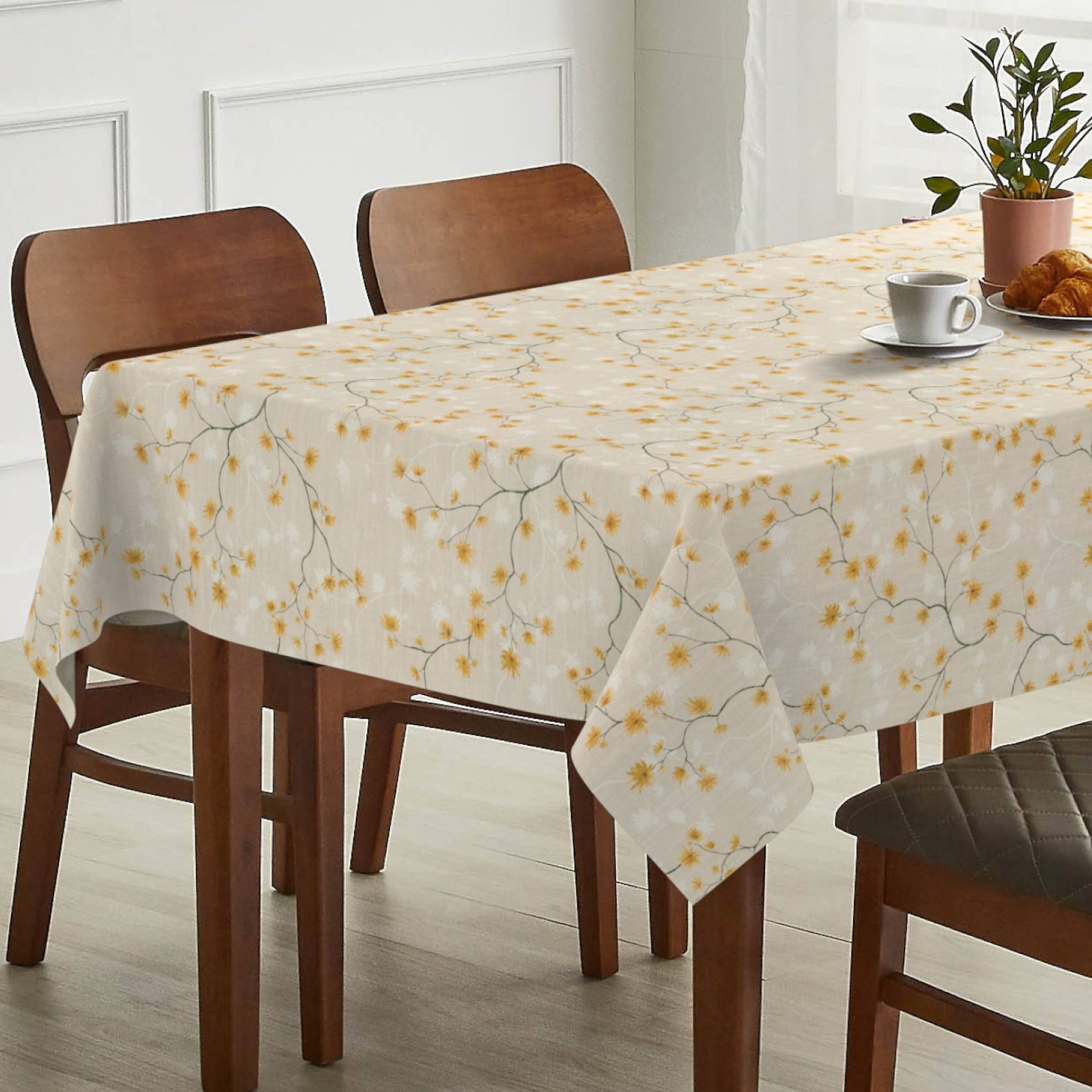 Esstisch mit weißer Blumentischdecke, Holzstühlen und einer Tasse, einem Teller, einem Croissant und einer Topfpflanze auf der Tischplatte.