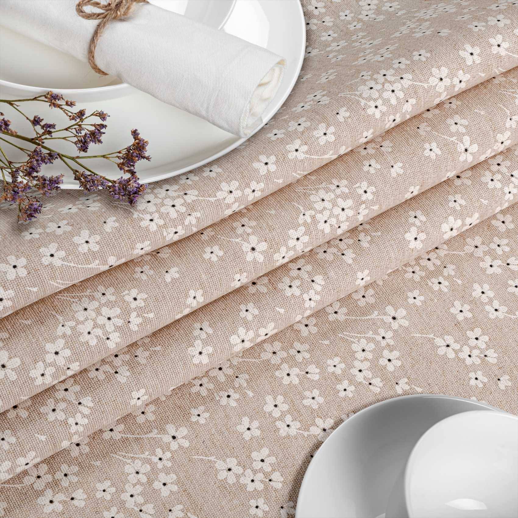 Beigefarbener Blumenstoff in diagonalen Falten neben einem Teller, zusammengerollter Serviette und getrockneten Blumen, der ein weiches Tischtuch-Textil zeigt.