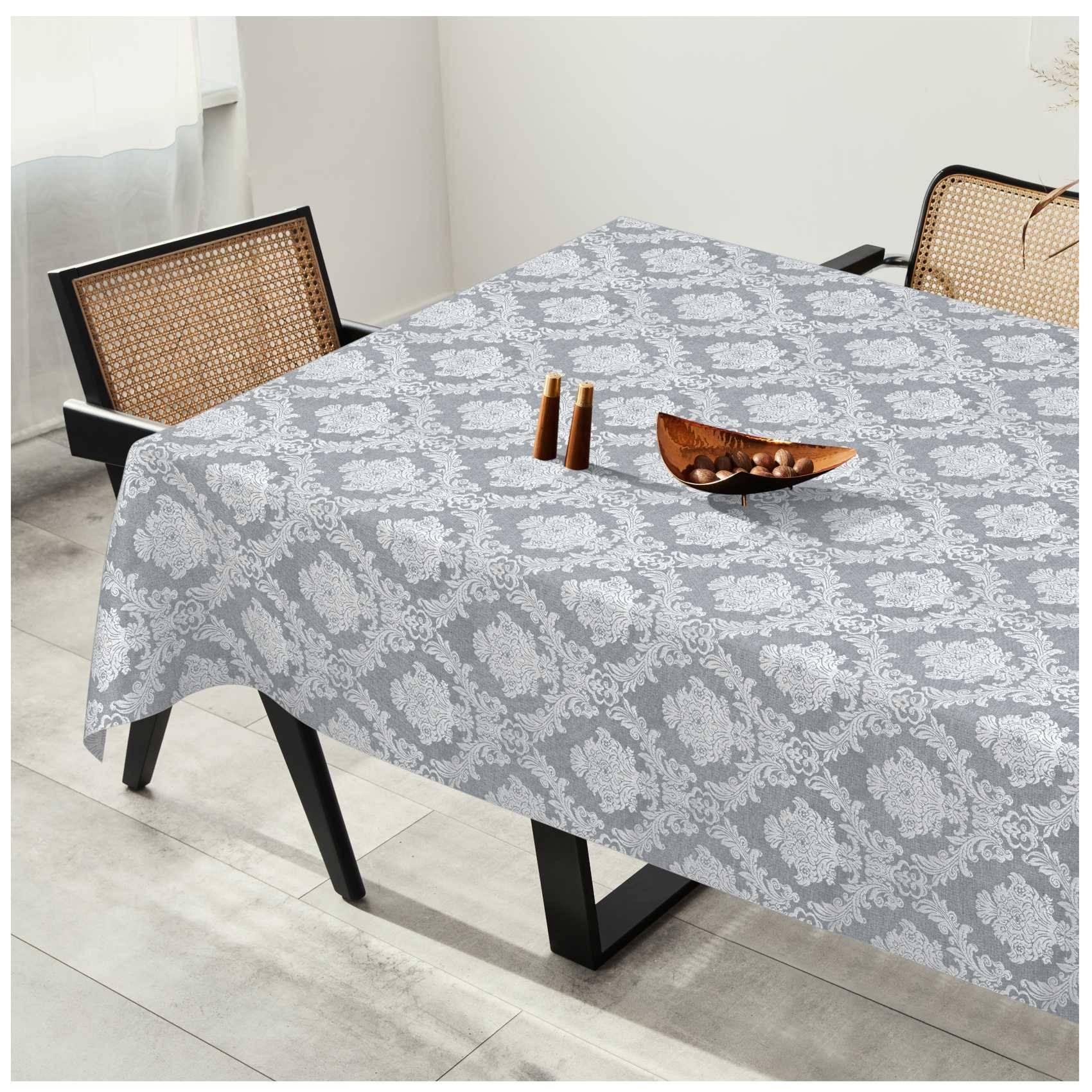 Esstisch mit einer grau gemusterten Tischdecke, eingedeckt mit zwei Kerzen und einer kleinen Holzschale, neben Stühlen mit gewebtem Rückenlehne in einem hellen Raum.
