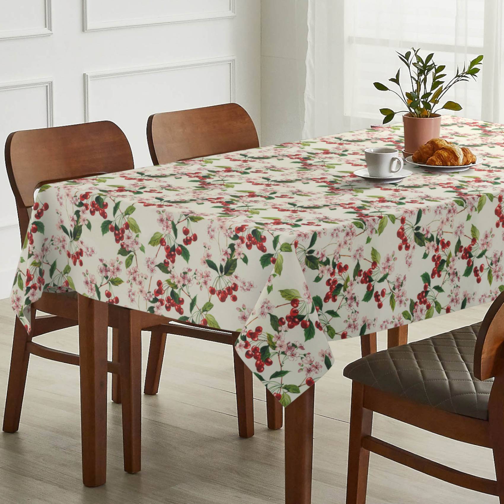 Esstisch mit floraler Tischdecke, eingedeckt mit einer Tasse, einem Croissant und einer Topfpflanze neben Holzstühlen in einem hellen Raum.