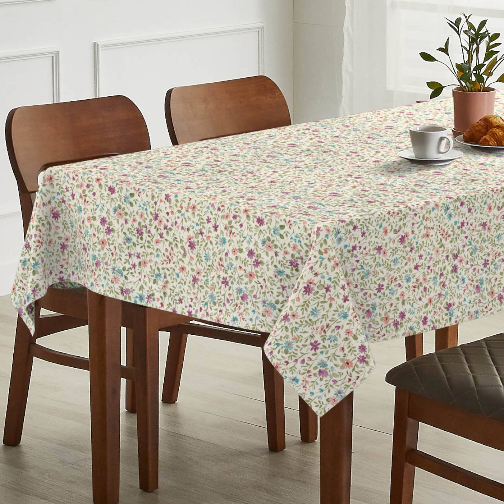 Esstisch mit floraler Tischdecke, Stühlen und einem Gedeck mit Tasse, Untertasse und Croissants in einem hellen Zimmer
