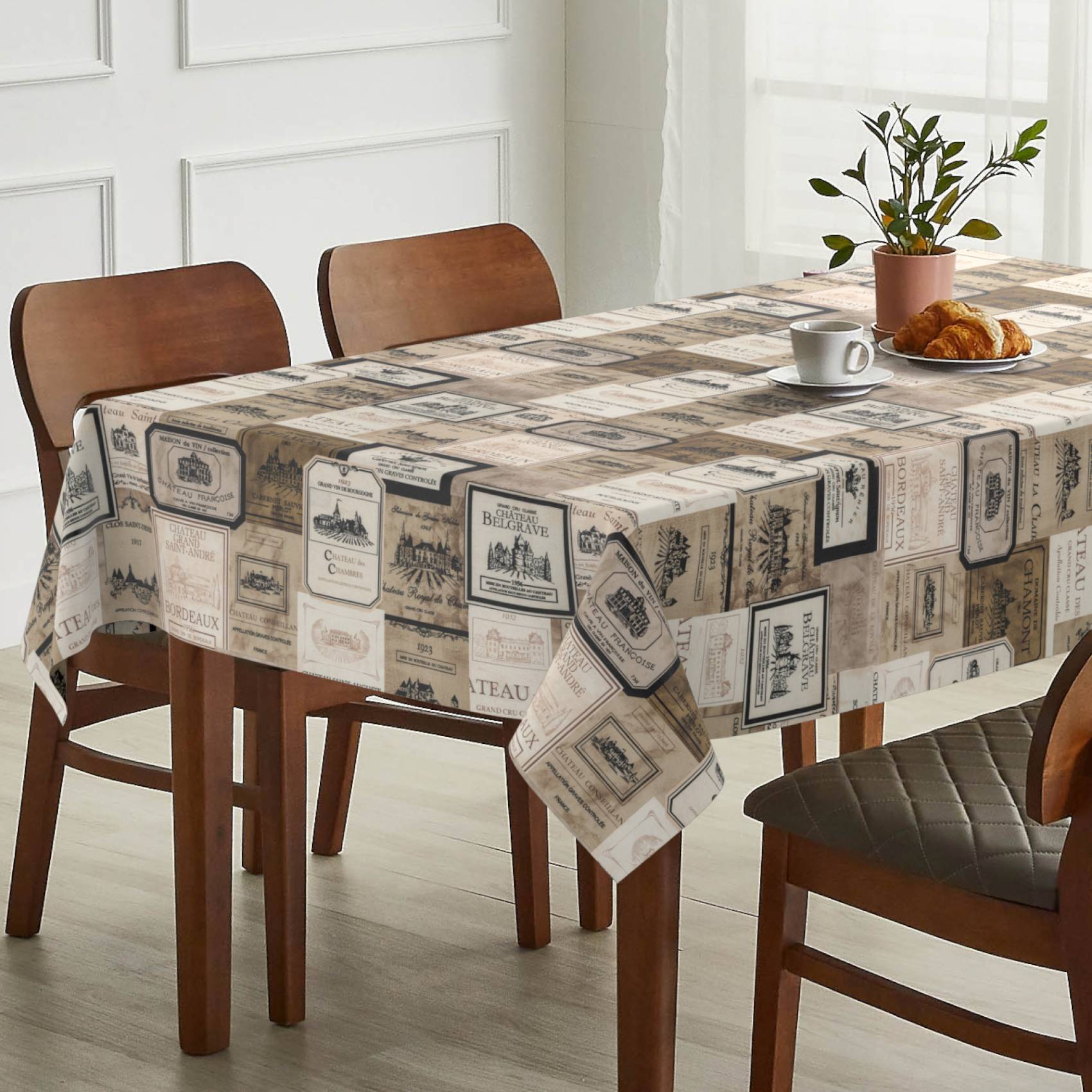 Esstisch mit einer Tischdecke im Vintage-Stil bedruckt, zwei Stühlen und Gedeck mit einer Kaffeetasse, Croissant und einer Topfpflanze neben einem hellen Fenster.