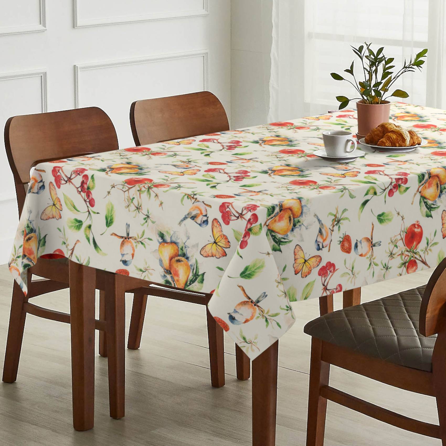 Esstisch mit floraler Schmetterlings-Tischdecke in einem hellen Raum, eingedeckt mit einer Pflanze, einer Kaffeetasse und einem Croissant auf einem Teller.