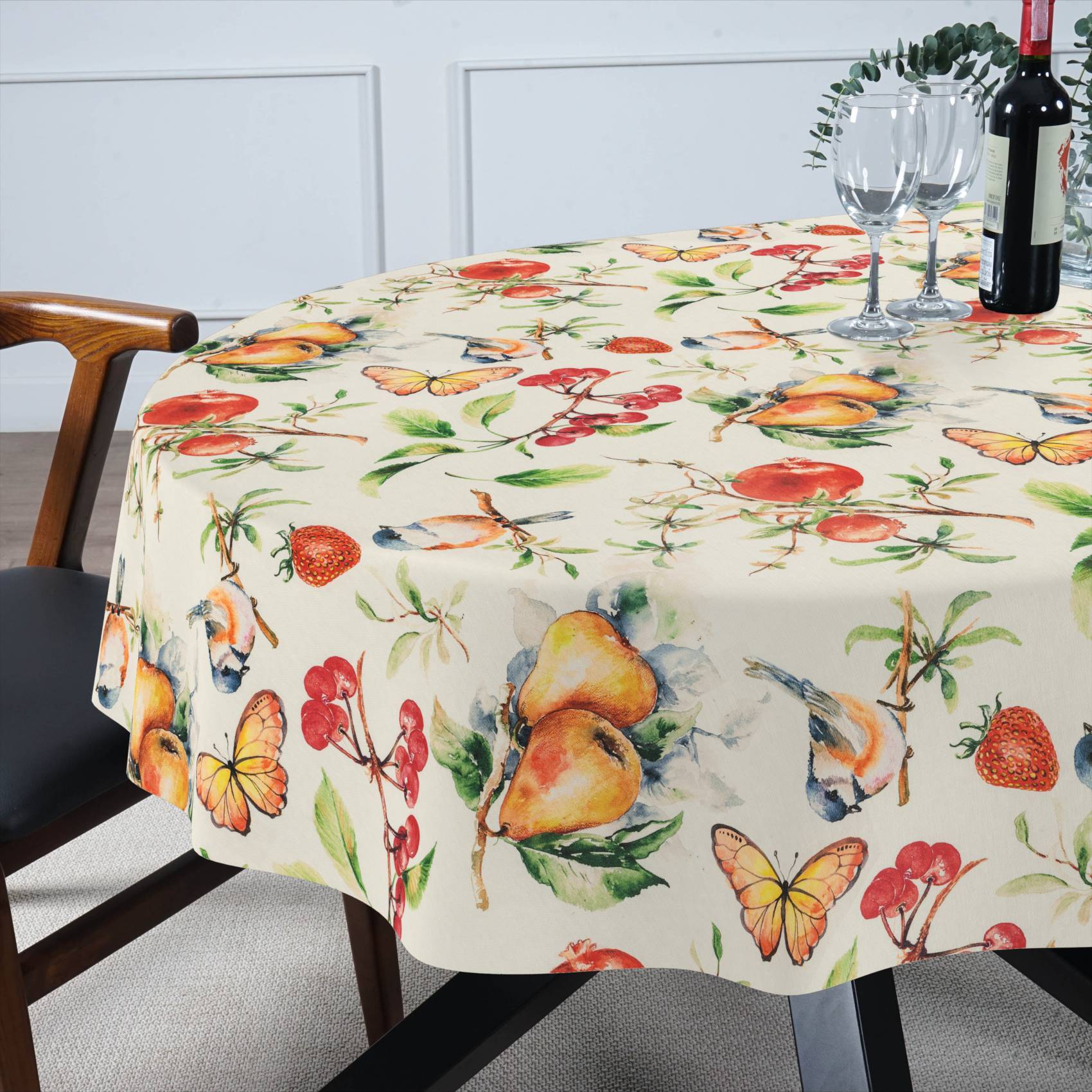 Runder Esstisch mit einer Tischdecke mit Obst- und Schmetterlingsmuster, eingedeckt mit Weingläsern und einer Flasche neben einem Stuhl.