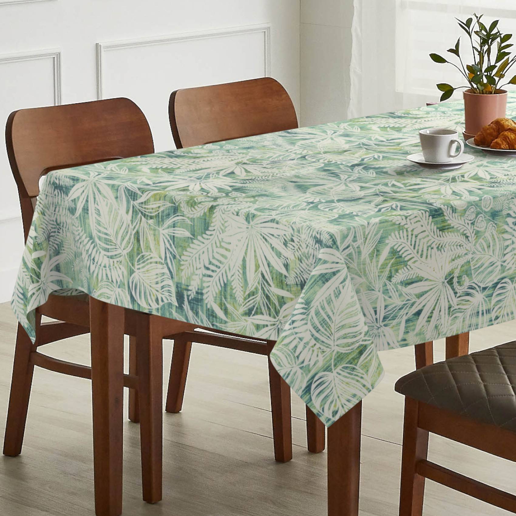 Esstisch mit einer grünen Tischdecke mit tropischem Blattmuster, eingedeckt mit einer Tasse, einem Croissant und einer Topfpflanze in einem hell erleuchteten Raum.