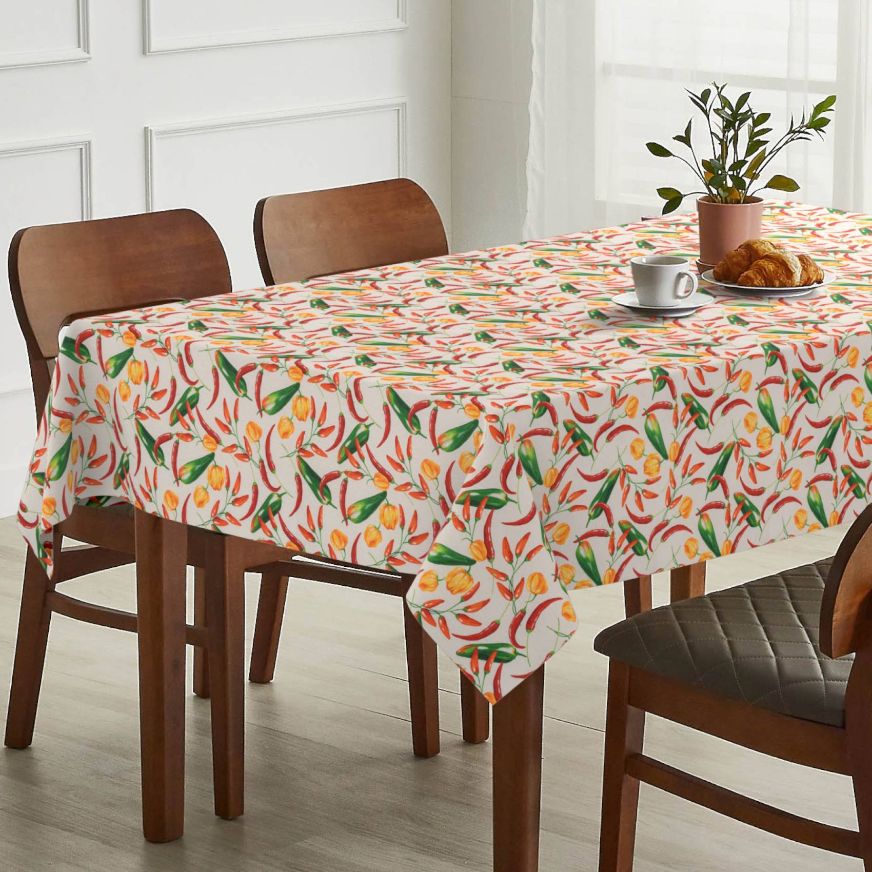 Esstisch mit farbenfroher fruchtgemusterter Tischdecke, eingedeckt mit Kaffee, Croissant und Topfpflanze in einem hellen Raum.