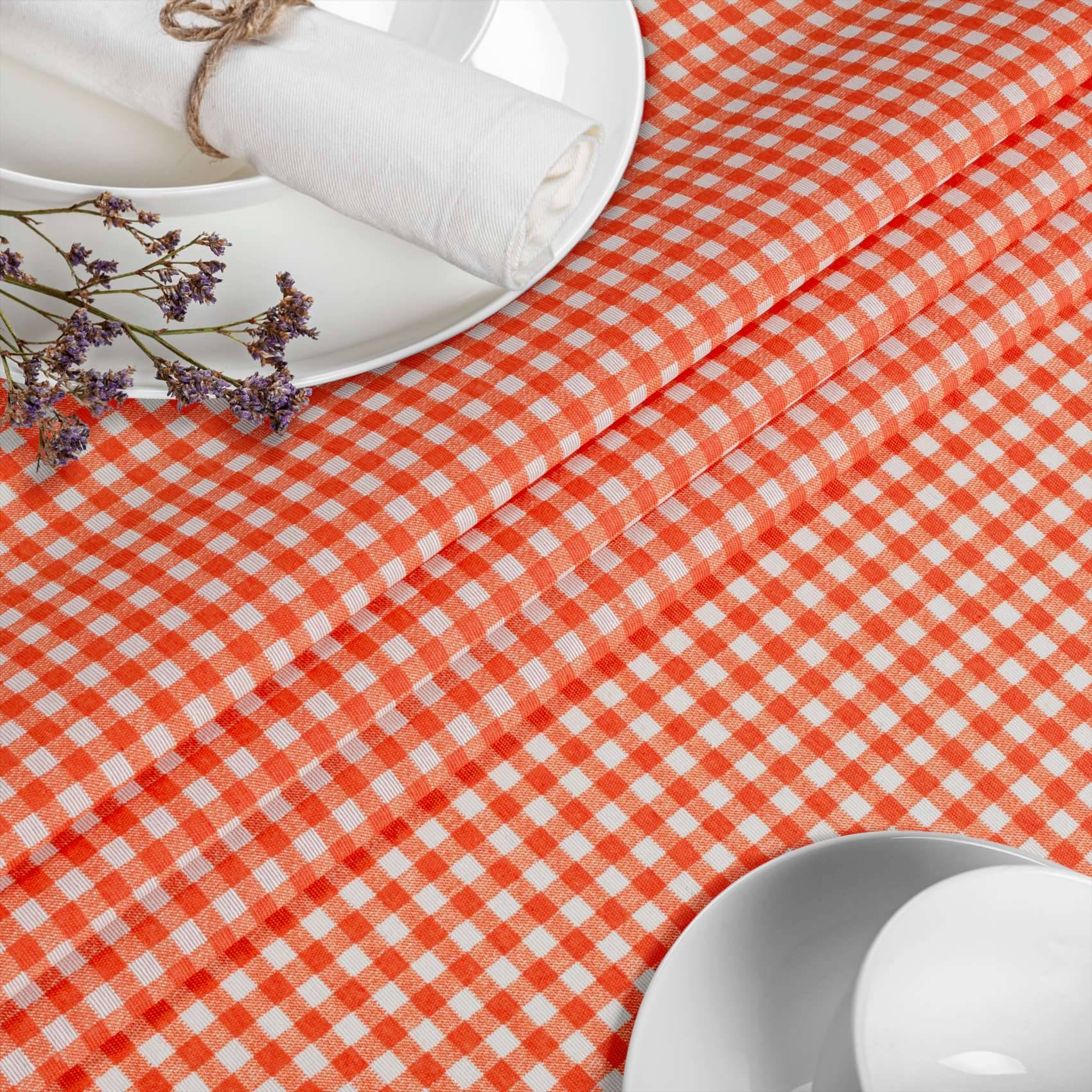 Weiß-orange kariertes Tischtuch mit stapelweise weißem Geschirr, gefalteter Serviette und getrockneten Blumen, was auf ein Picknick oder eine Tischdeckung hindeutet.