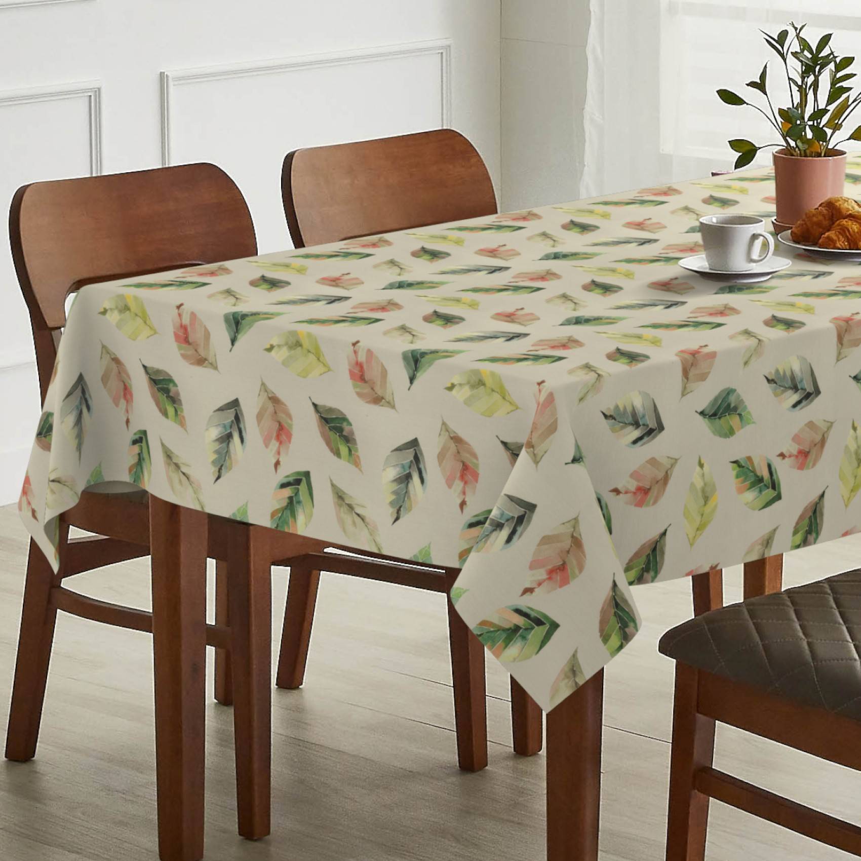 Esstisch mit blattgemusterter Tischdecke, Stühlen und einer Tasse, einem Teller, einem Croissant und einer Topfpflanze in einem hellen Raum.