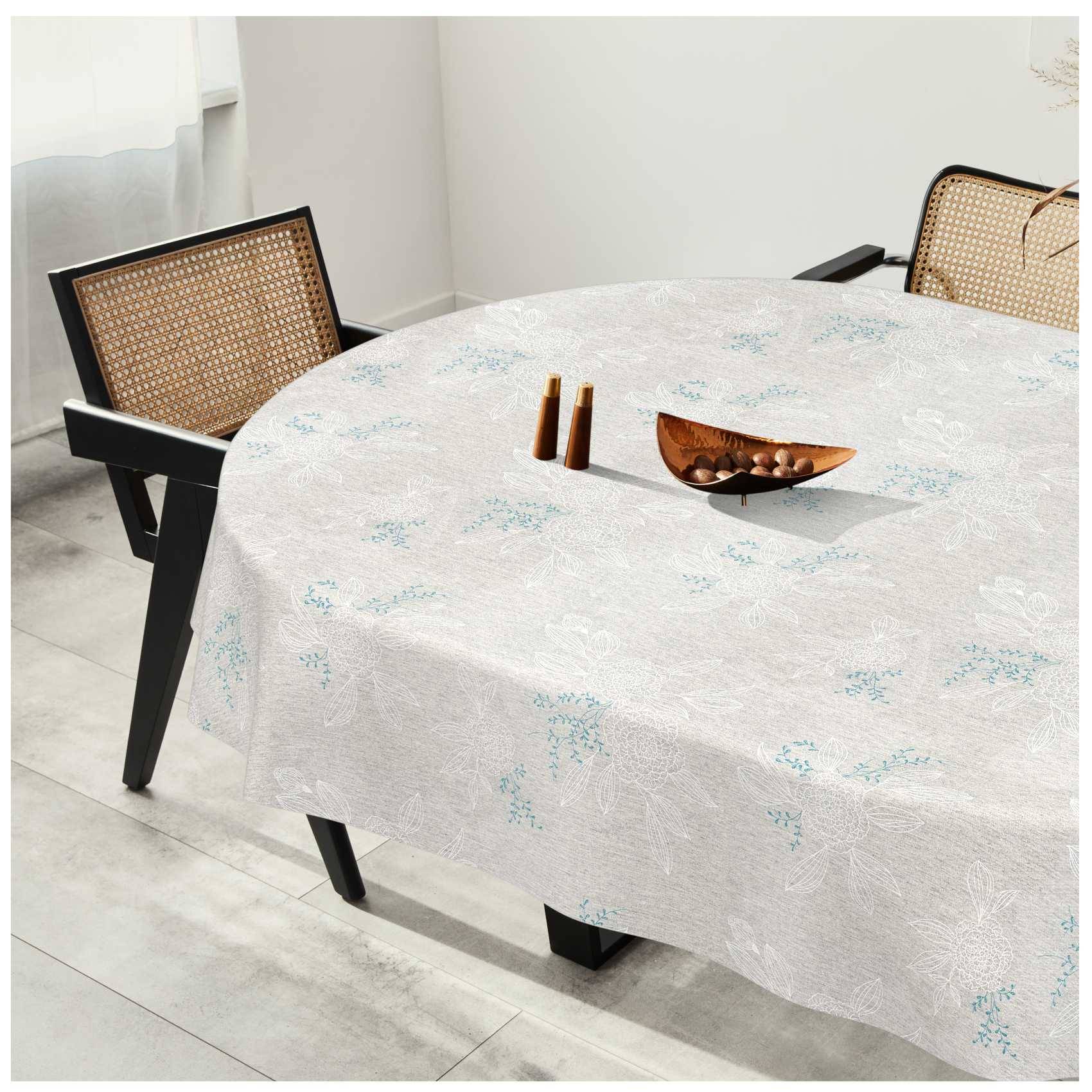 Esstisch mit einer leicht gemusterten Tischdecke, zwei geflochtenen Stühlen und einer Holzschüssel mit zwei kleinen Flaschen darauf in einem hellen Zimmer.