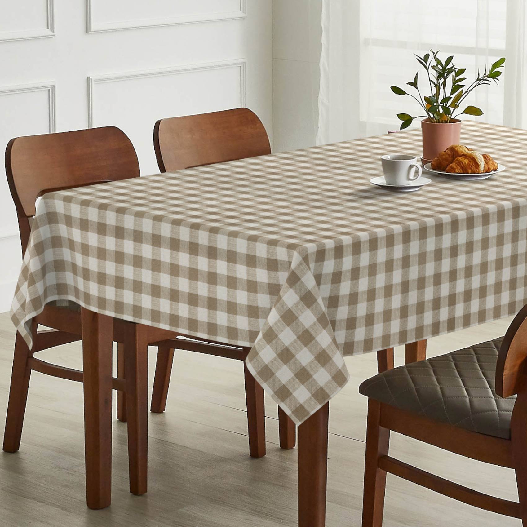 Esstisch mit beigem Karomuster-Tischdecke, vier Holzstühlen und einer Kaffeetasse, einem Croissant und einer Topfpflanze darauf in einem hellen Raum.