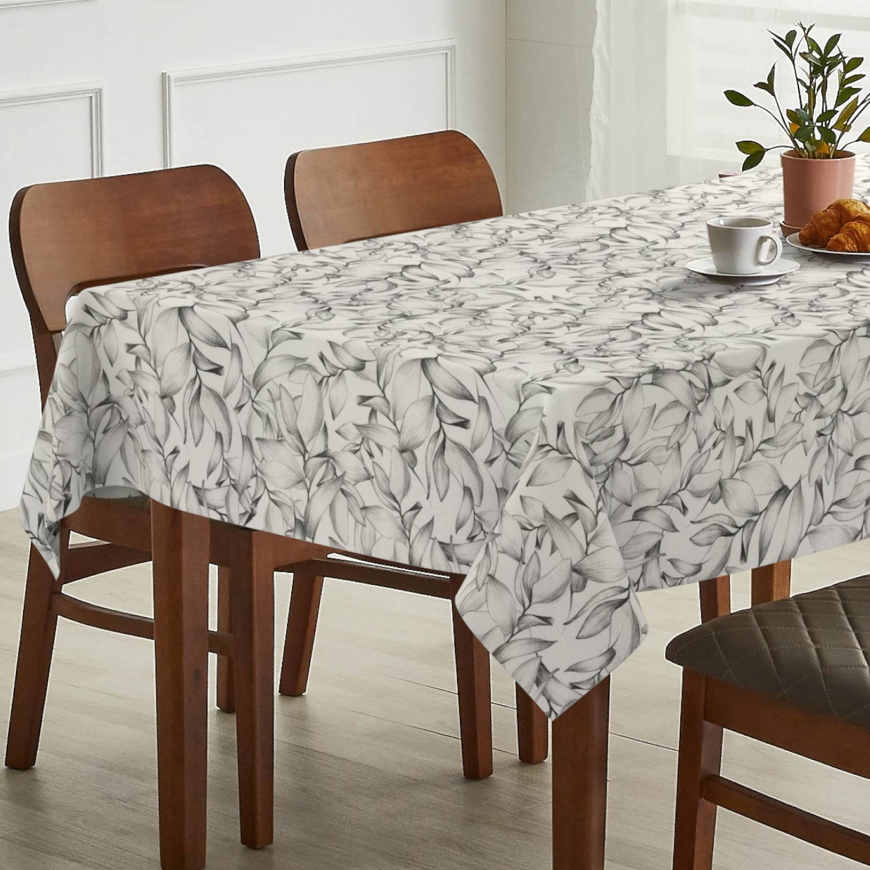Esstisch mit einer blattgemusterten Tischdecke, mit Stühlen, einer Tasse und einem Croissant in einem hellen Raum.