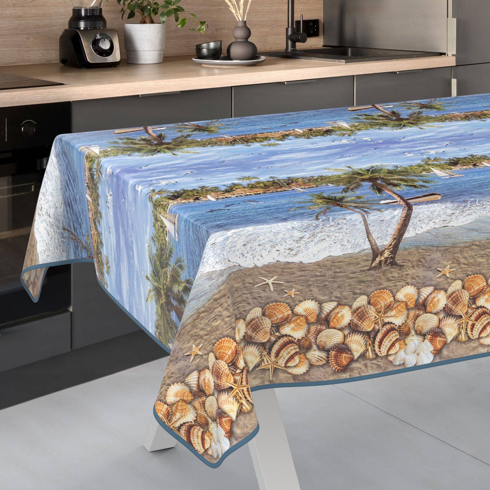 Tischdecke mit Strandszenen-Druck, die Ozean, Sand, Muscheln und Treibholz auf einem Esstisch in einer modernen Küche zeigt.
