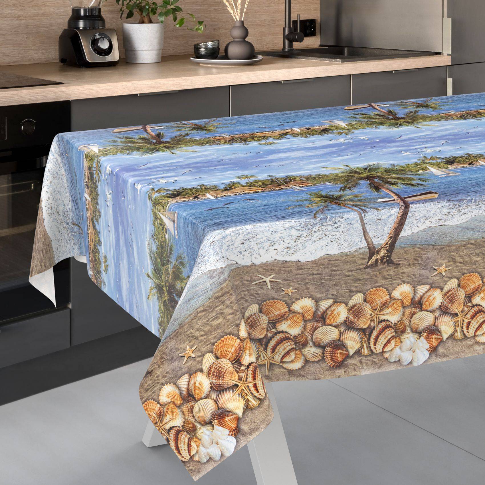 Küchentisch mit einer strandthematischen Tischdecke bedeckt, bedruckt mit Muscheln, Sand, Wasser und Treibholz, über eine moderne Küchentheke drapiert.